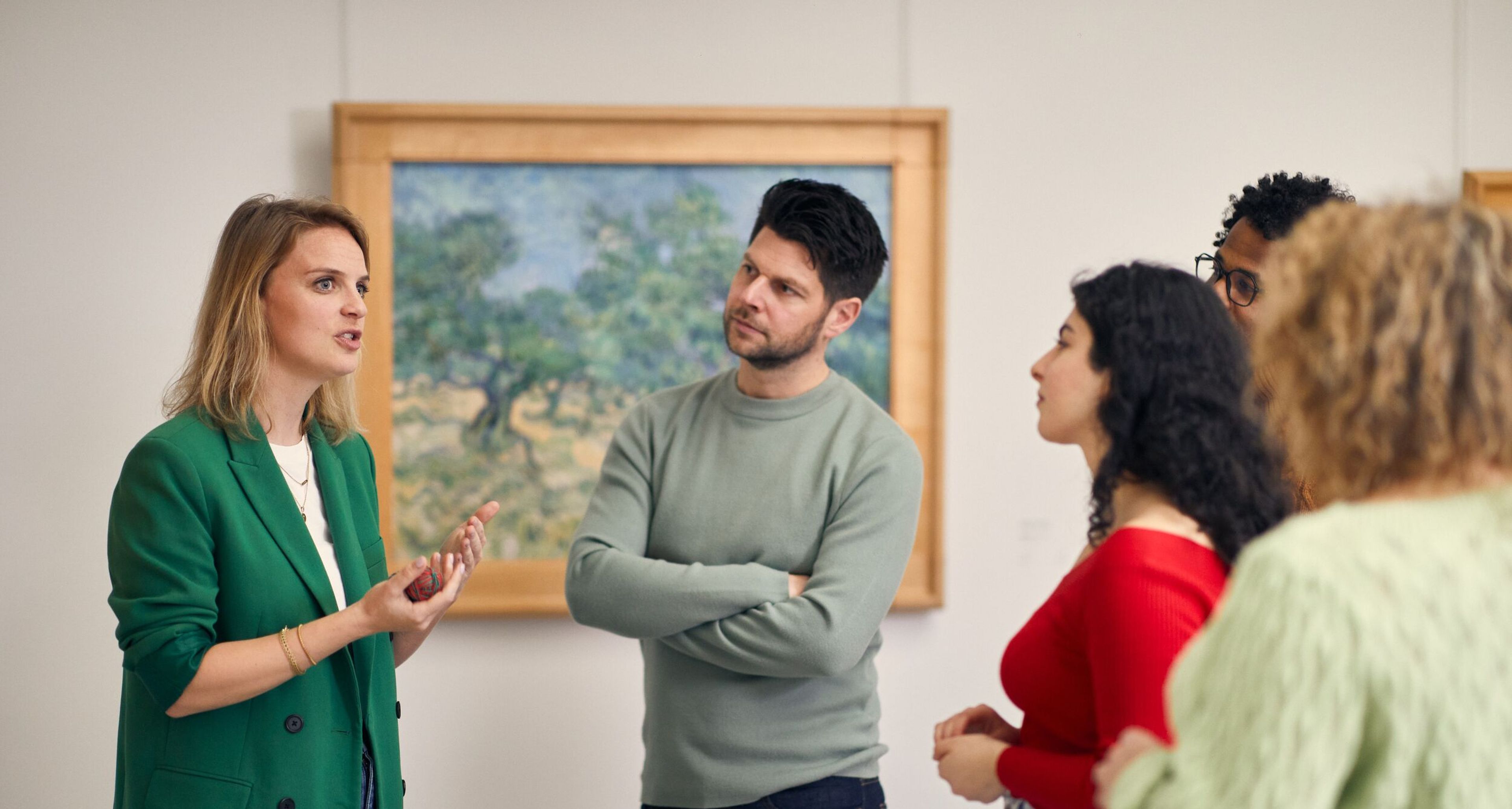 Rondleider vertelt aan een groep met mensen, in de Van Gogh galerij. Op de achtergrond een schilderij van Van Gogh met olijfbomen, in groen, blauw en geel.