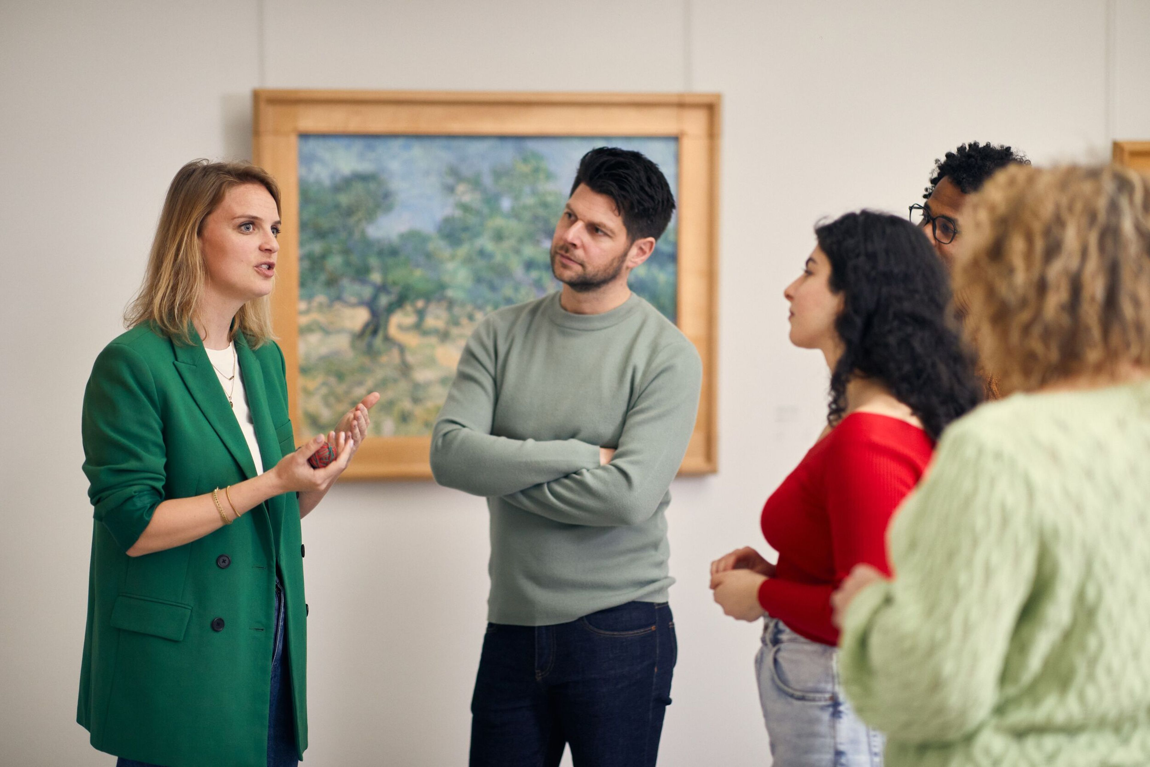 Rondleider vertelt aan een groep met mensen, in de Van Gogh galerij. Op de achtergrond een schilderij van Van Gogh met olijfbomen, in groen, blauw en geel.