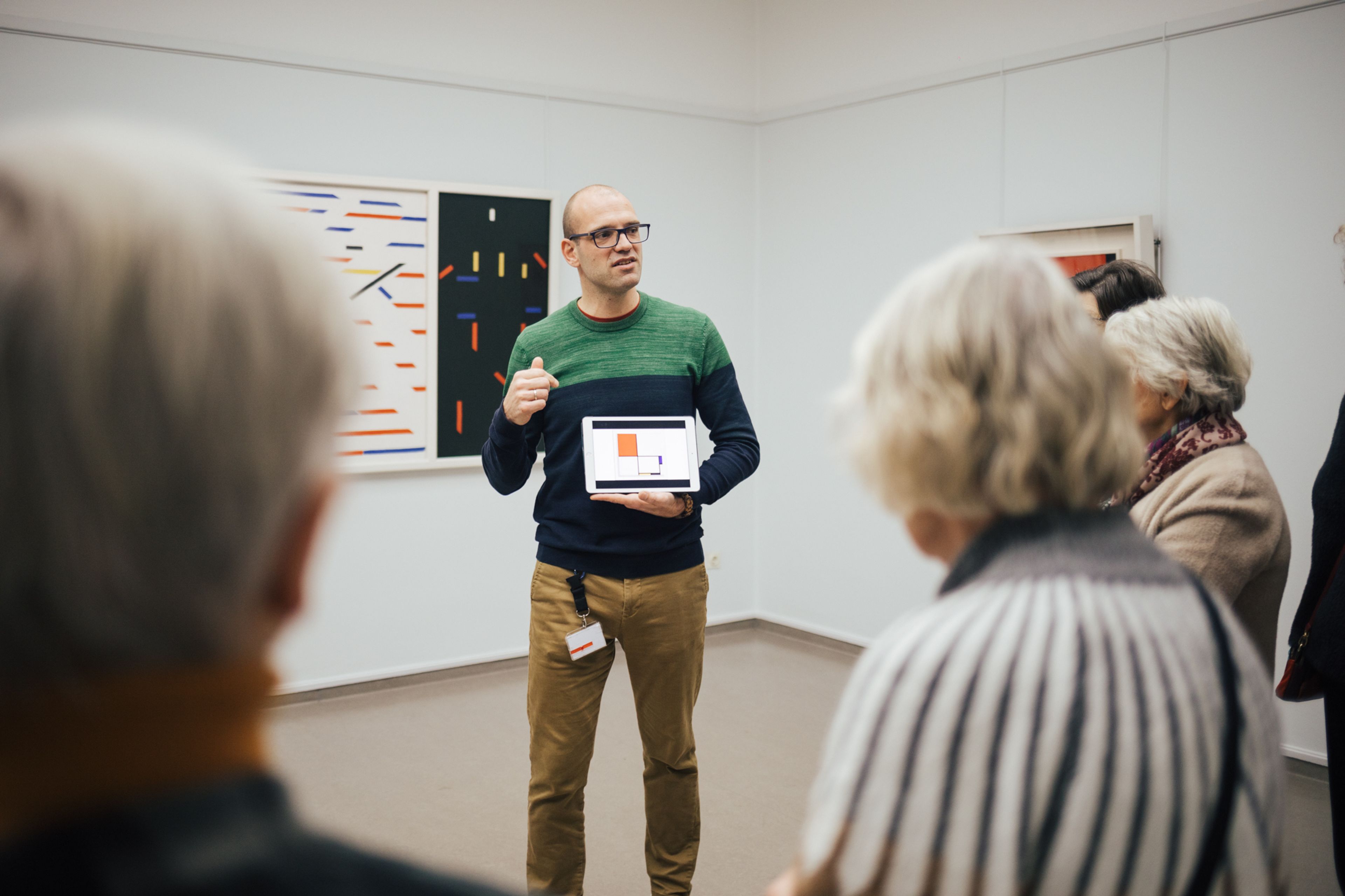 Foto van een man die een rondleiding geeft aan een kleine groep oudere volwassenen in een galerijruimte met moderne kunst aan muren. Man toont een tablet met een grafiek, waarbij aandacht van publiek gericht is op uitleg over kunstwerken en gegevens.