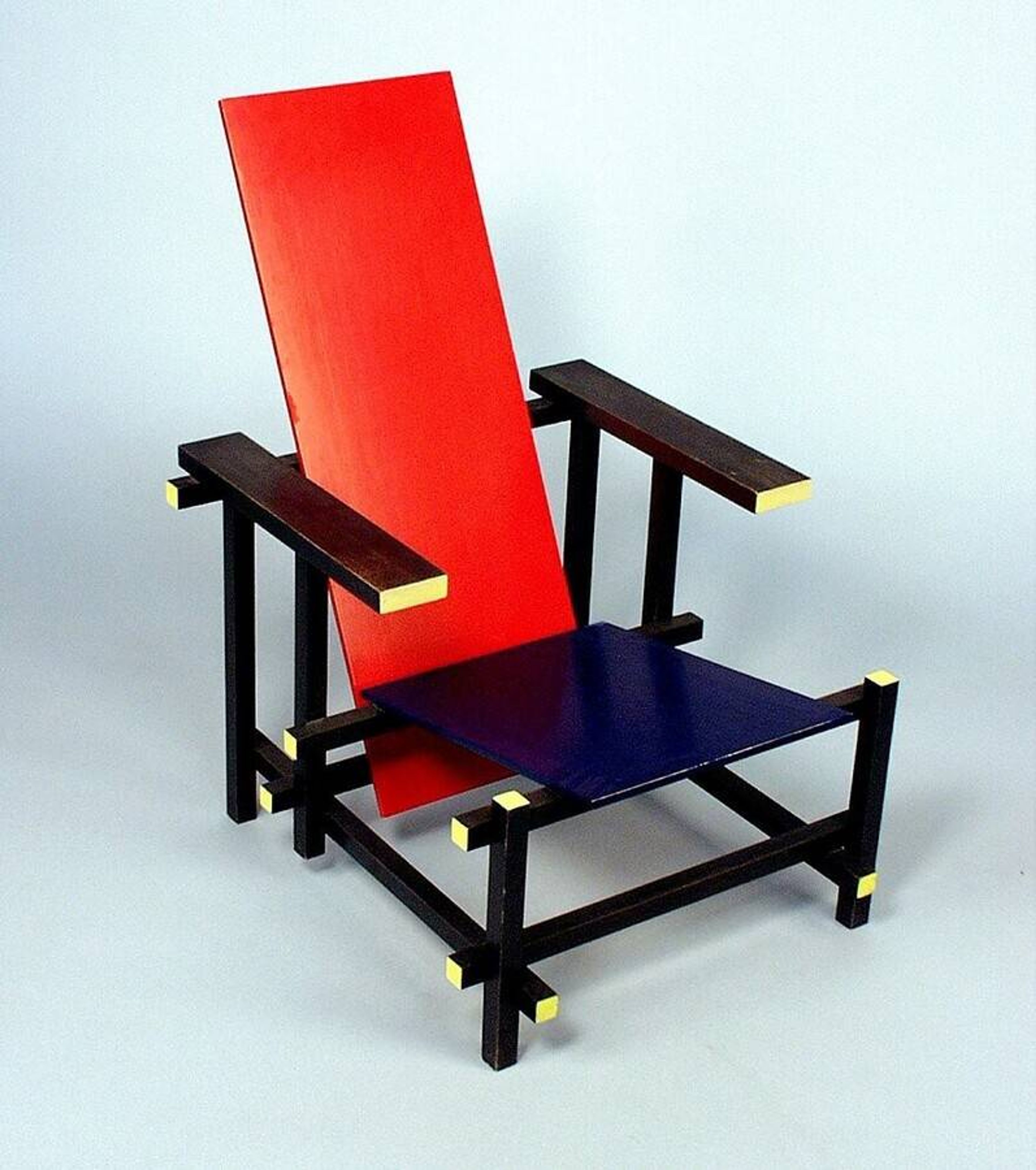 Gerrit Rietveld, stoel, ontwerp 1918, latere uitvoering Foto van een moderne stoel met vlakken, bestaande uit een rood rugleuningpaneel, een blauwe zitting en een zwart frame met gele accenten op hoeken. Ontwerp benadrukt rechte lijnen en contrasterende kleuren, typerend voor De Stijl kunstbeweging.
