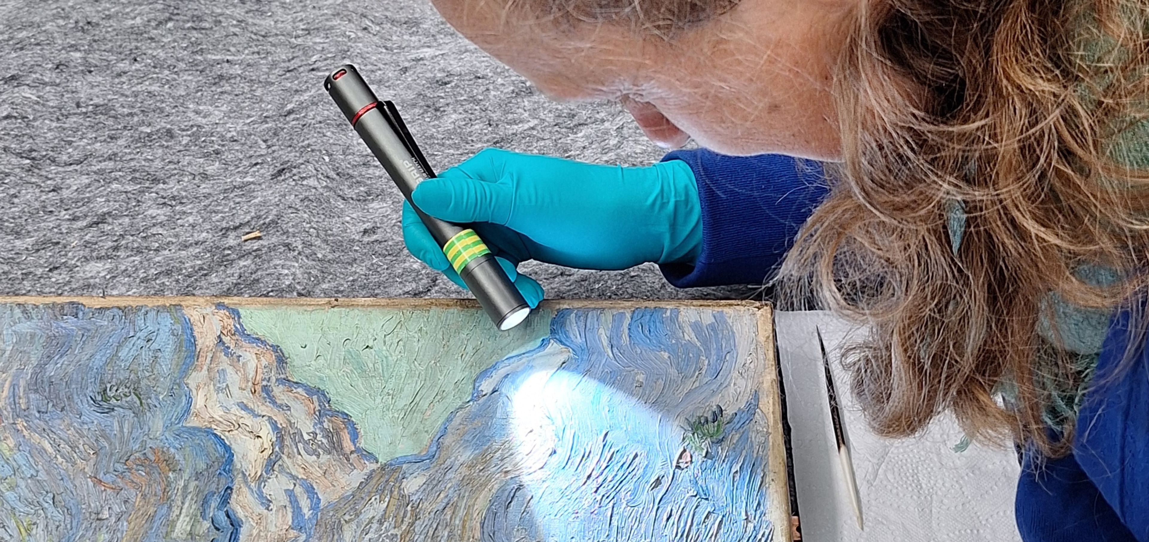Foto van iemand die op een atelier aandachtig met een zaklamp kijkt naar een schilderij (Van Gogh)