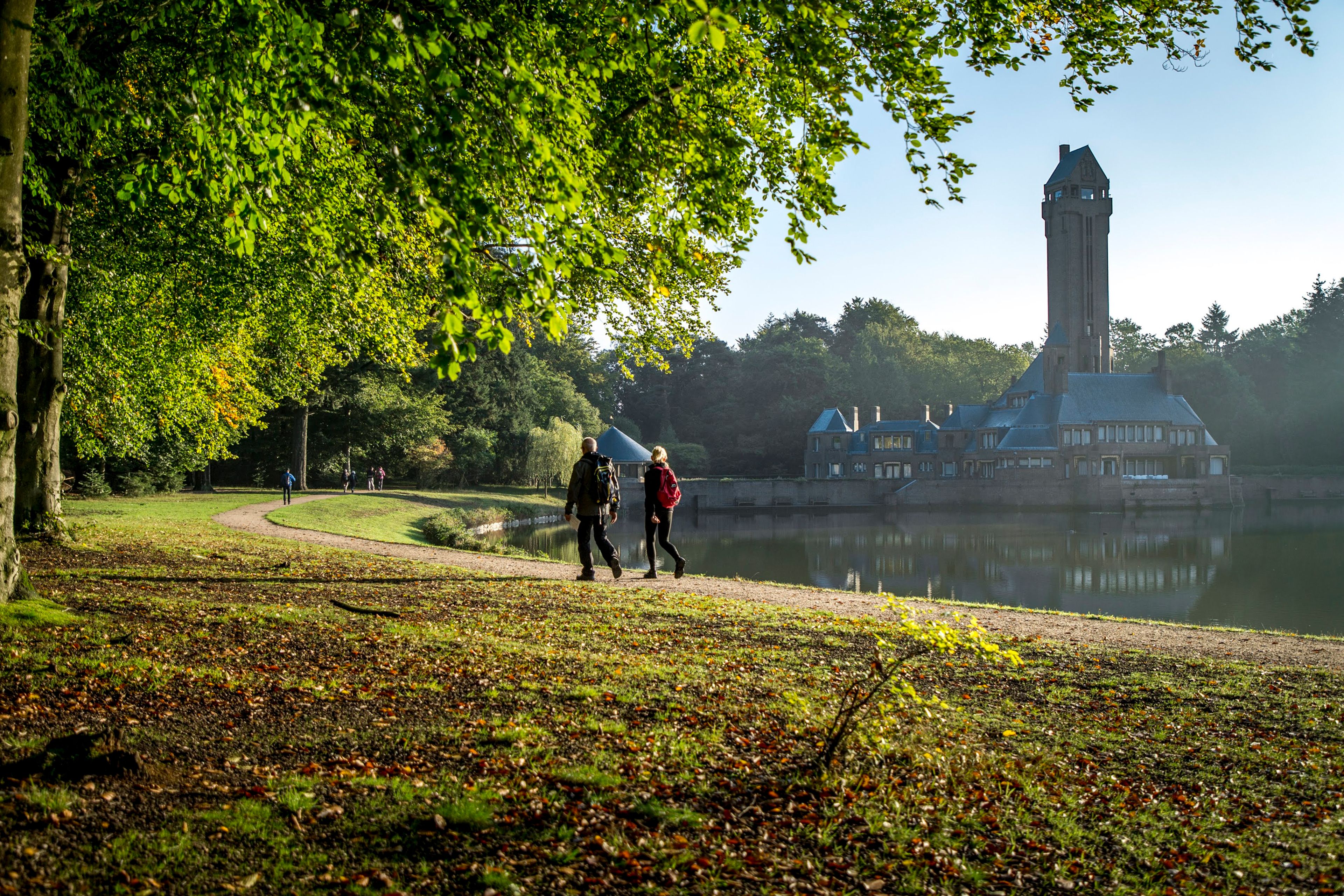 Foto van twee wandelaars langs een waterkant en Jachthuis Sint Hubertus, een groot gebouw met hoge toren op de achtergrond. Zonlicht valt door groene bladeren