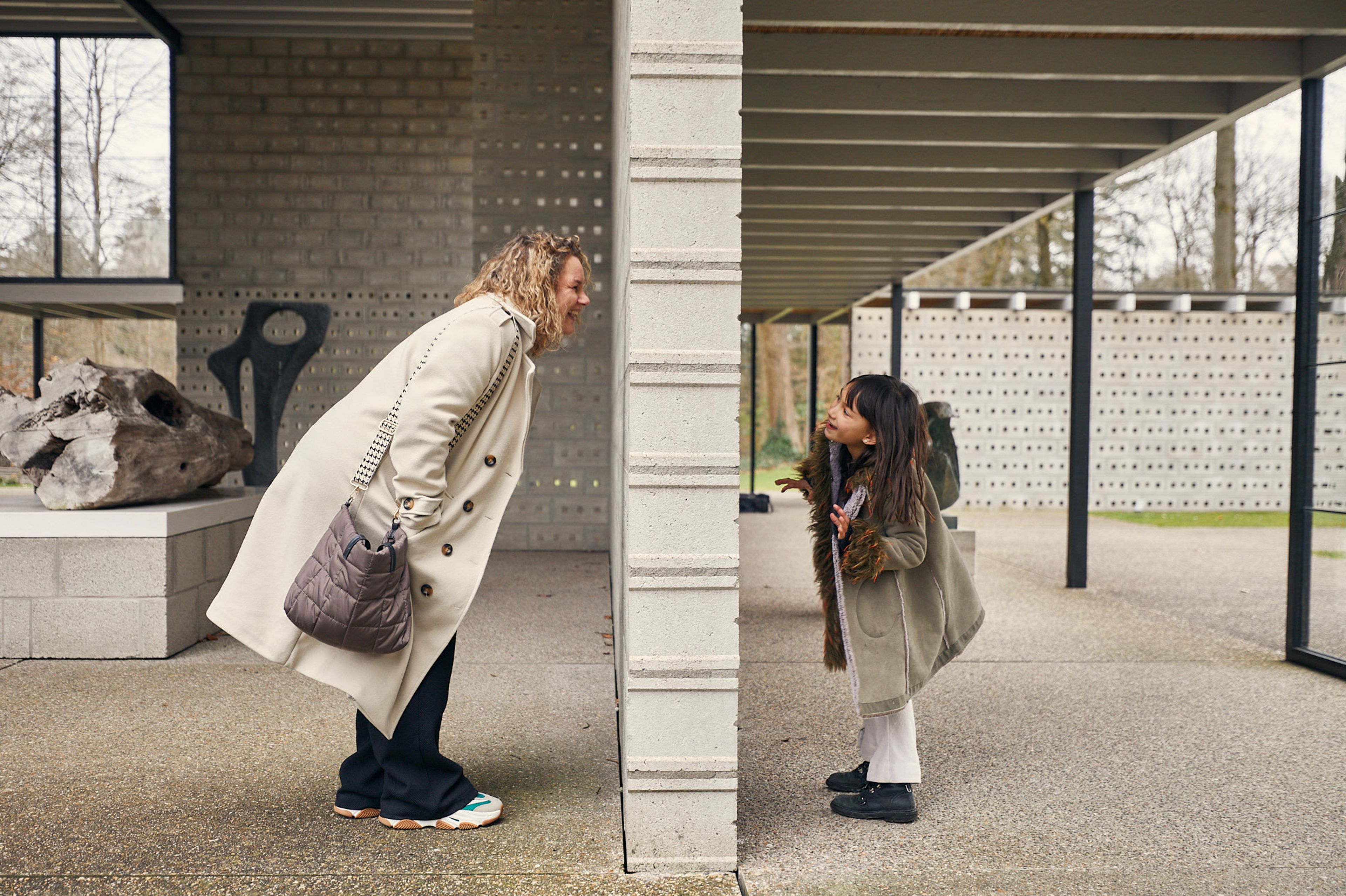 Fotografie van een vrouw en een meisje die aan weerszijden van een betonnen muur naar elkaar kijken, met een moderne, open architectuur op de achtergrond. Vrouw draagt een beige jas en draagt een grijze tas, meisje heeft lang haar en draagt een groene jas met witte broek.