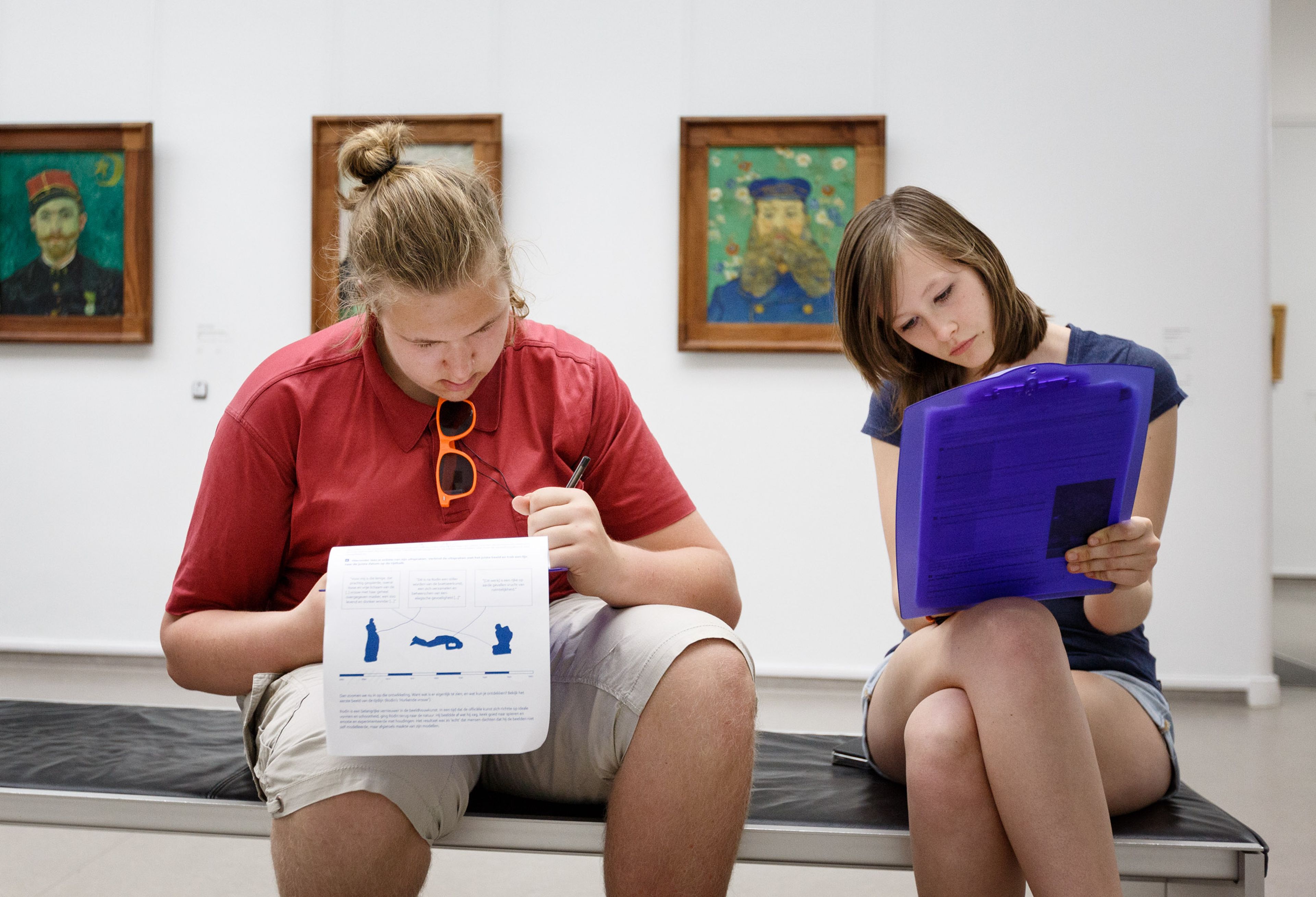Two people are sitting on a bench in a museum room with three paintings on the wall behind them. They study documents, with one person holding a sheet with blue images and the other a blue folder.