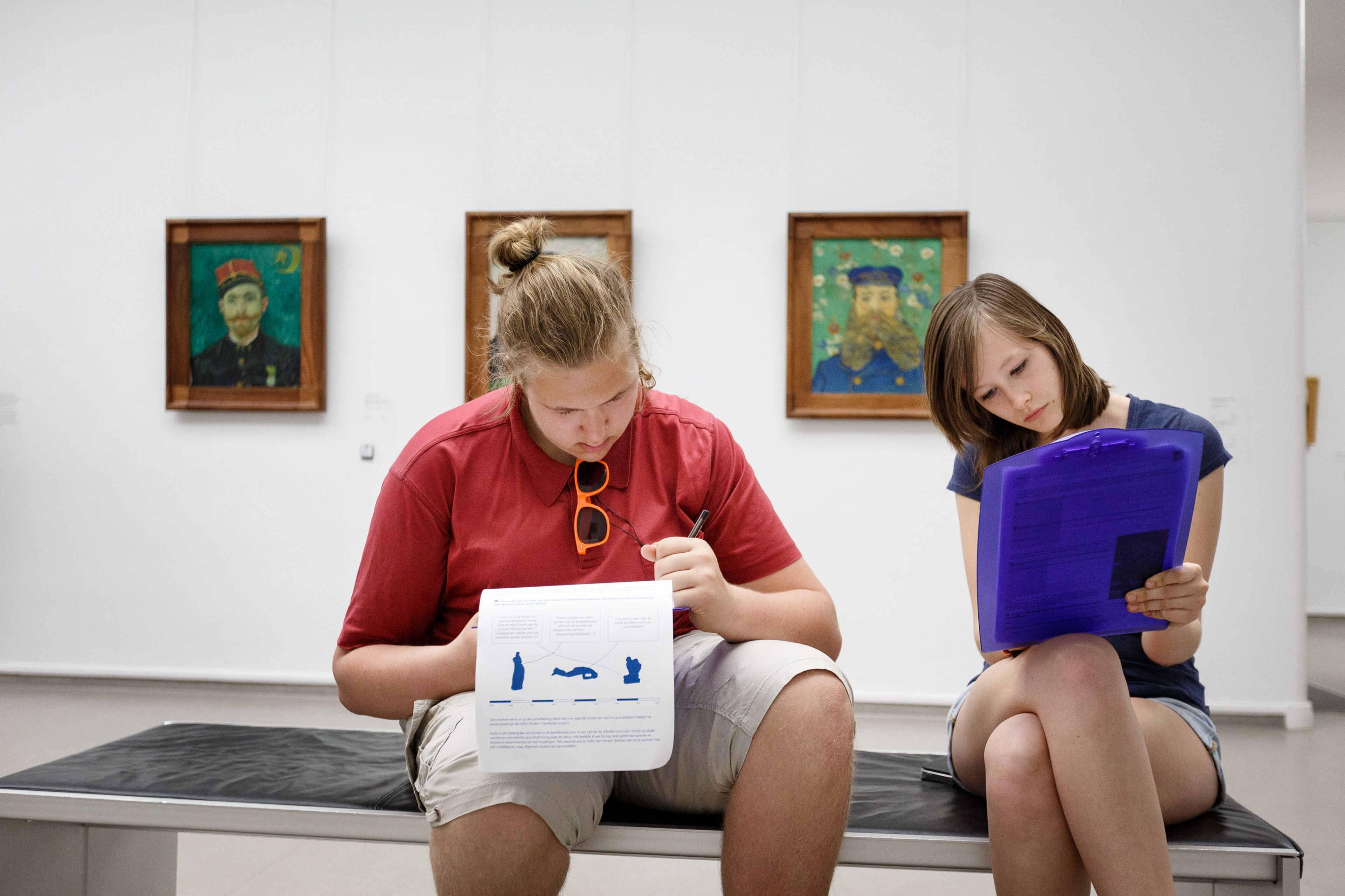 Twee personen zitten op een bank in een museumzaal met drie schilderijen aan de muur achter hen. Ze bestuderen documenten, waarbij één persoon een blad met blauwe afbeeldingen vasthoudt en de ander een blauw mapje.