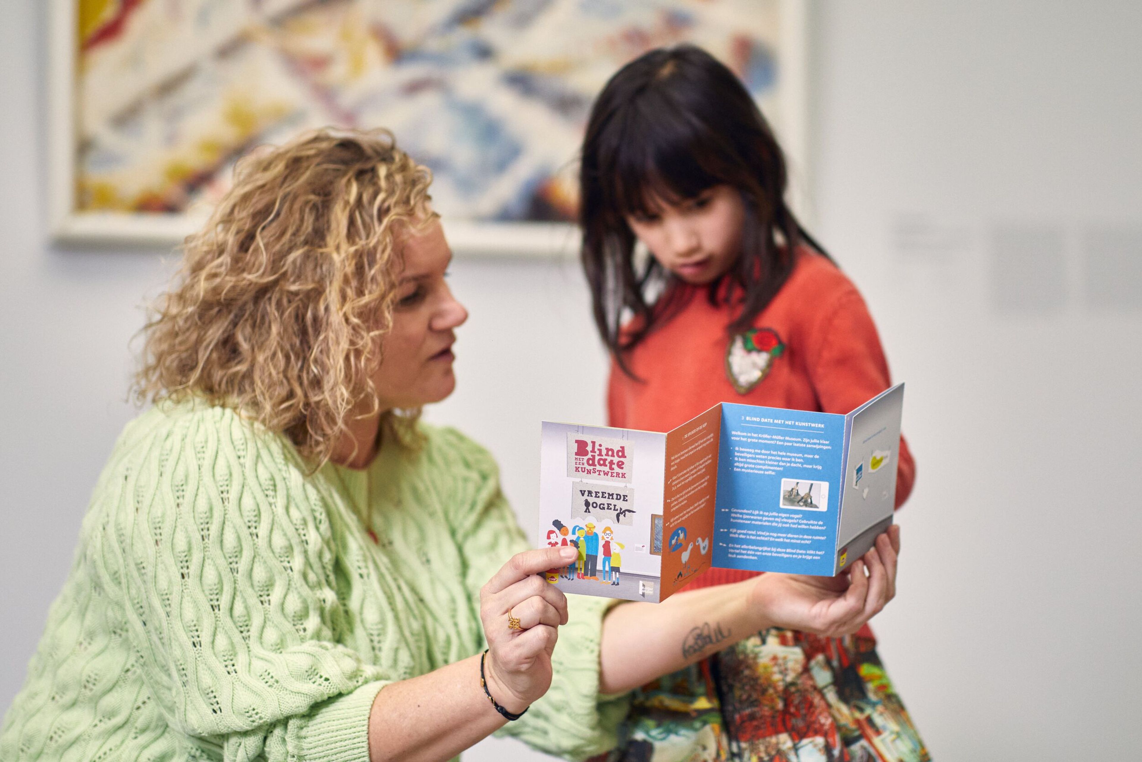 Foto van een vrouw en een kind die samen een boekje bekijken in een museum. Vrouw draagt een groene trui, kind een rode trui met een kleurrijke rok. Op de achtergrond hangt een abstract kunstwerk aan de muur.
