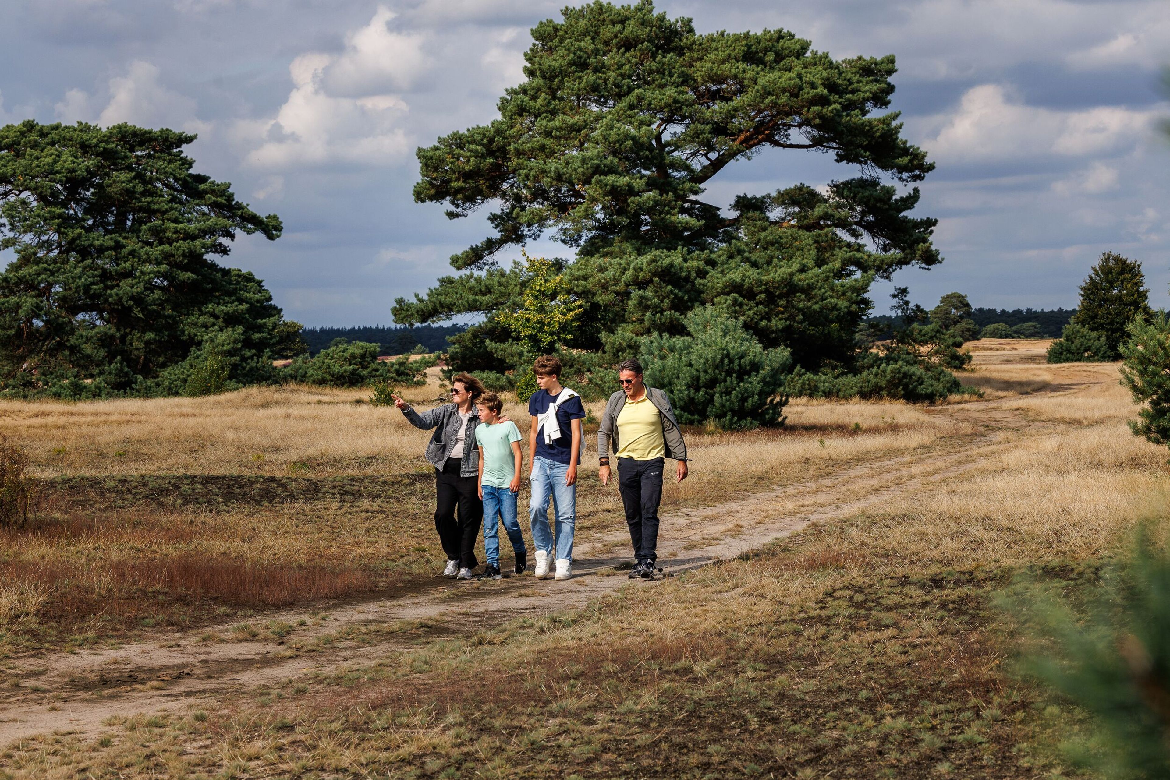 Twee volwassenen en twee kinderen wandelen over het gras, met hoge bomen op de achtergrond.