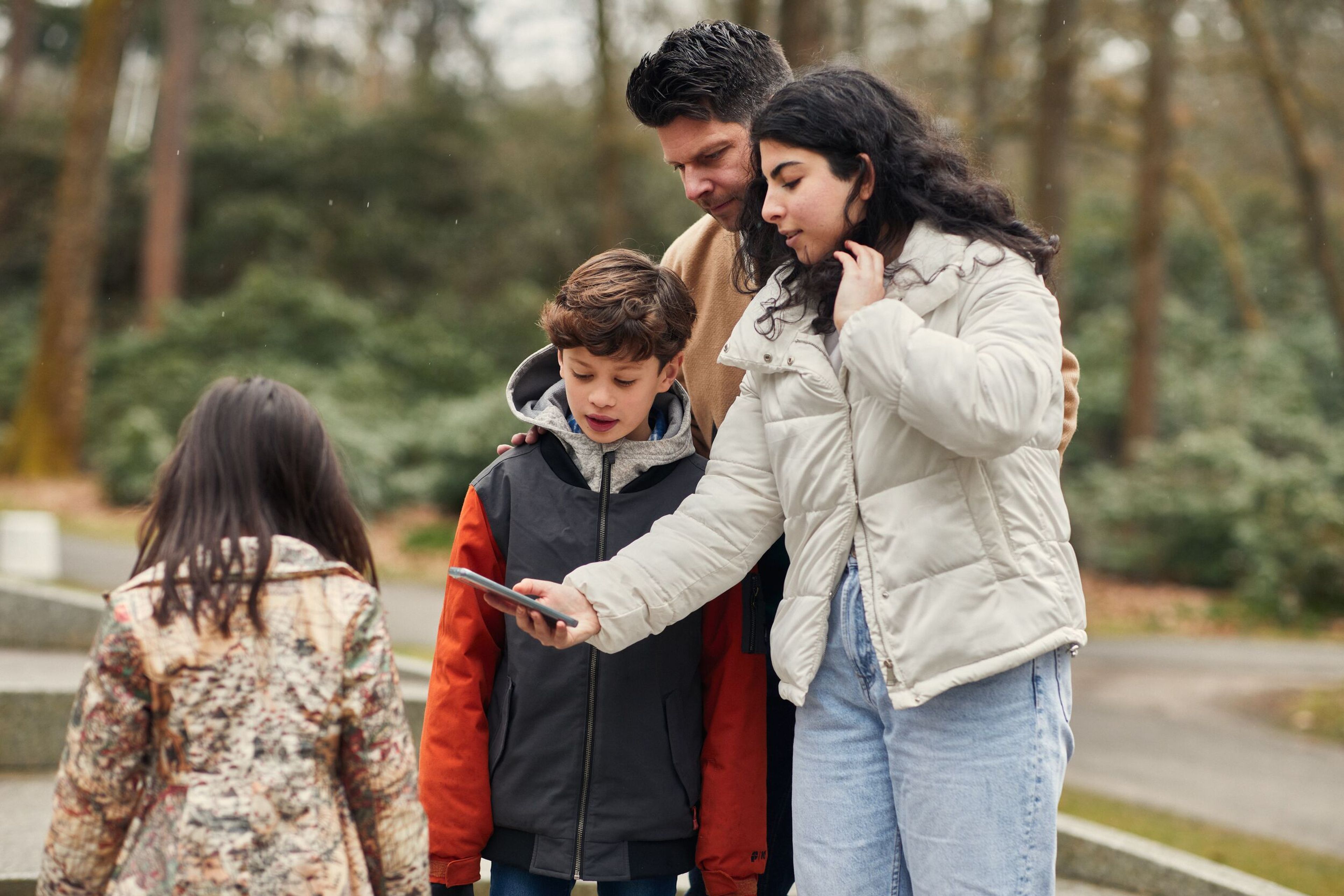 Foto van een groep van vier personen in een park, waarbij één persoon een smartphone vasthoudt en deze aan de anderen laat zien. De groep is gekleed in winterkleding en wordt omringd door bomen en groen.