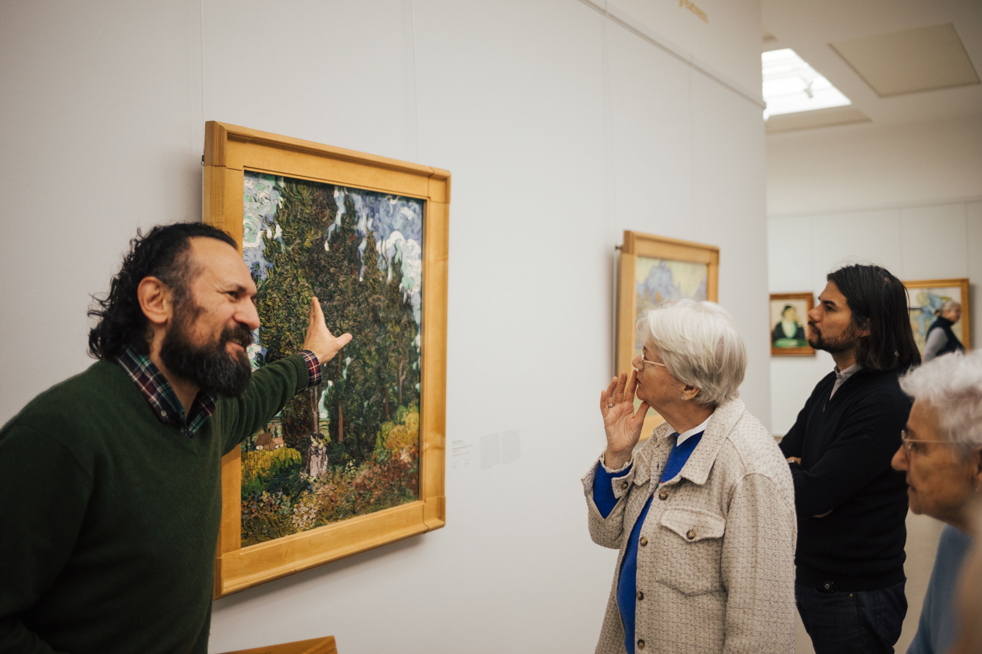 Foto van een groep mensen die naar een schilderij in een museum kijken, waarbij een man het kunstwerk aanwijst en uitlegt. Schilderij toont een boslandschap met groene en gele tinten, omgeven door een houten lijst, terwijl bezoekers aandachtig luisteren en observeren.