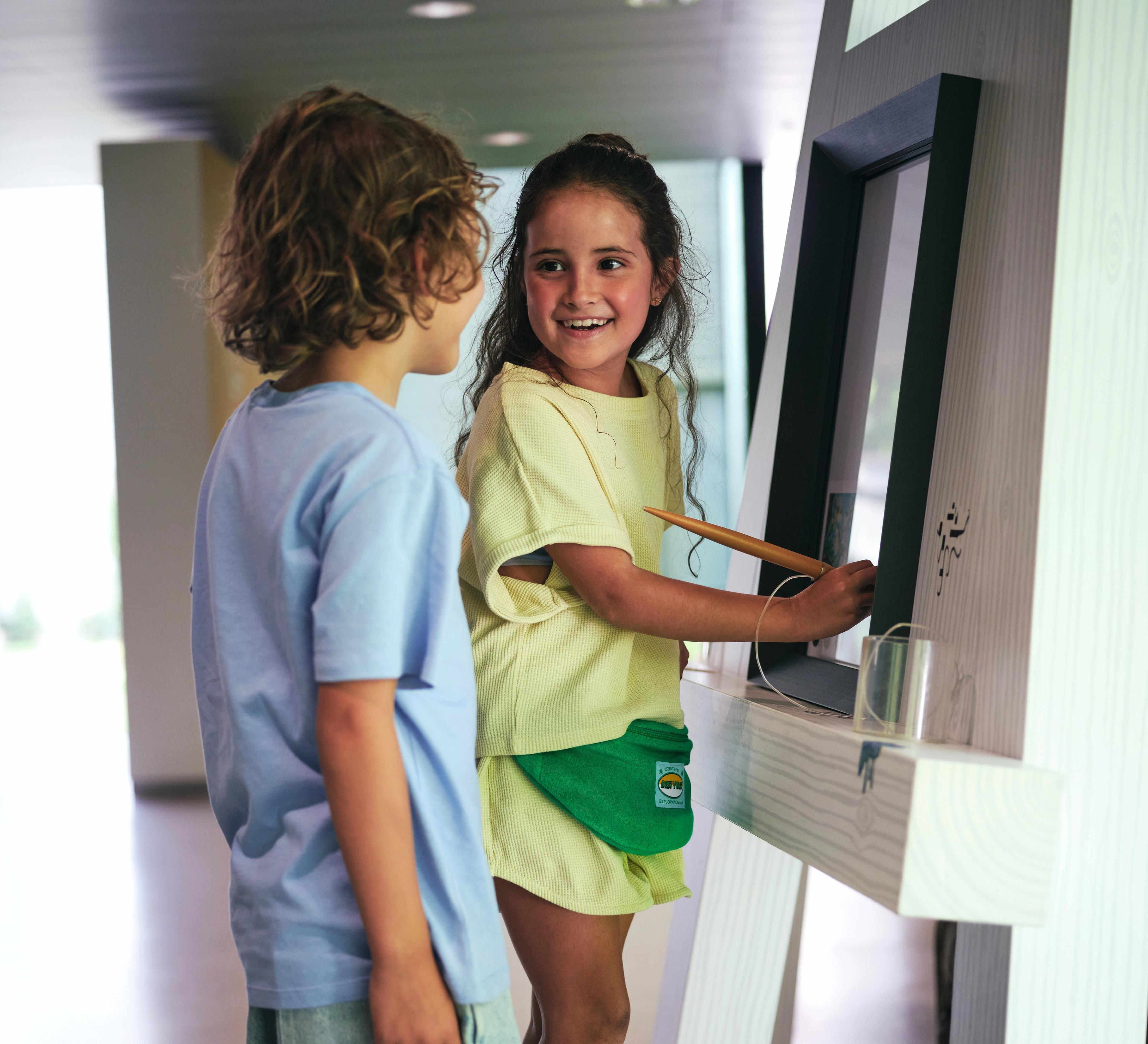 Foto van twee kinderen bij een interactieve schildersezel waarbij een meisje het scherm bedient en een jongen toekijkt. De schildersezel heeft een modern ontwerp met een witte standaard en een groot scherm, wat wijst op educatief of informatief gebruik in een openbare ruimte.