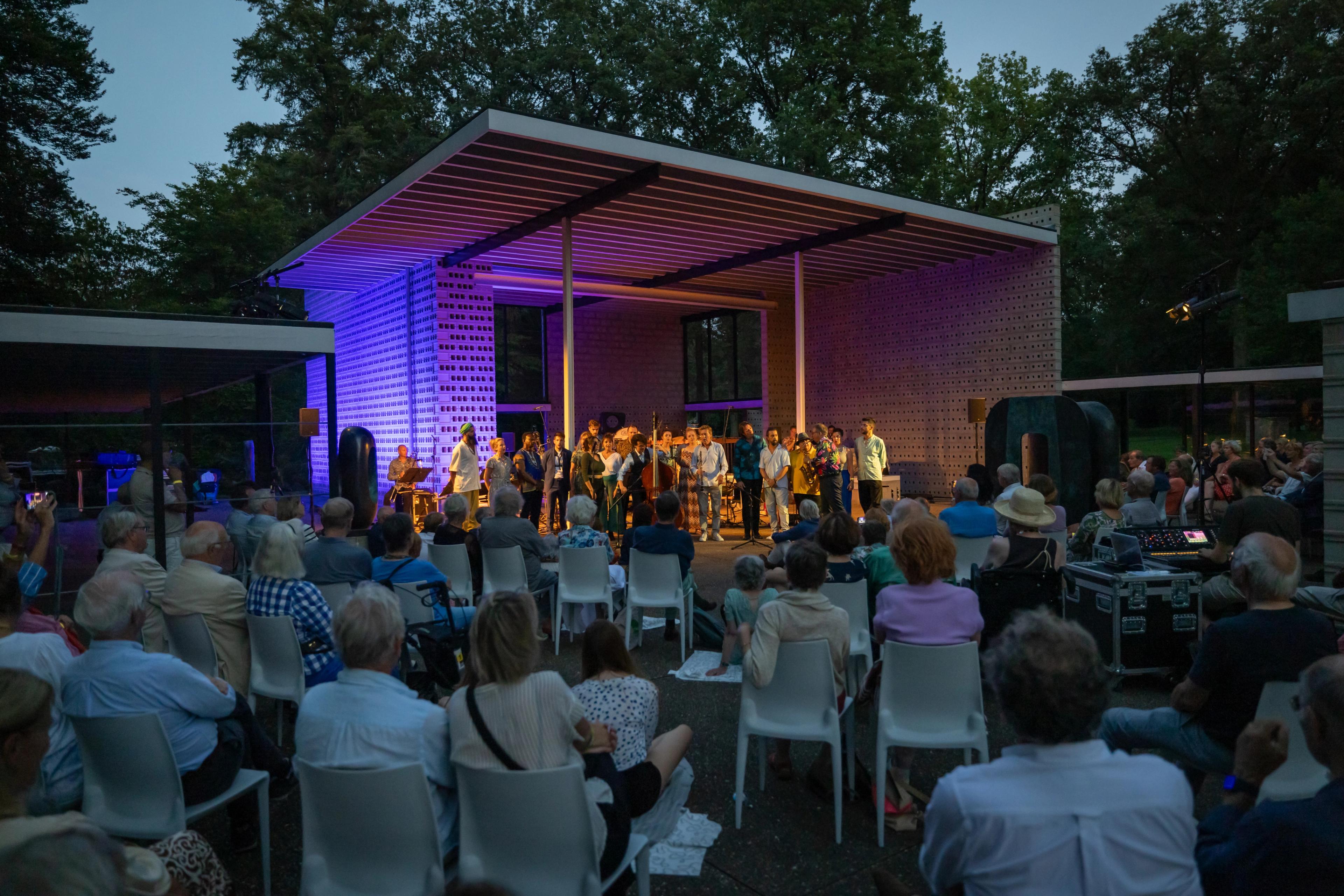 Foto van een modern paviljoen waar een optreden is, publiek staat en zit te kijken. Het is avond, het paviljoen is blauw verlicht.