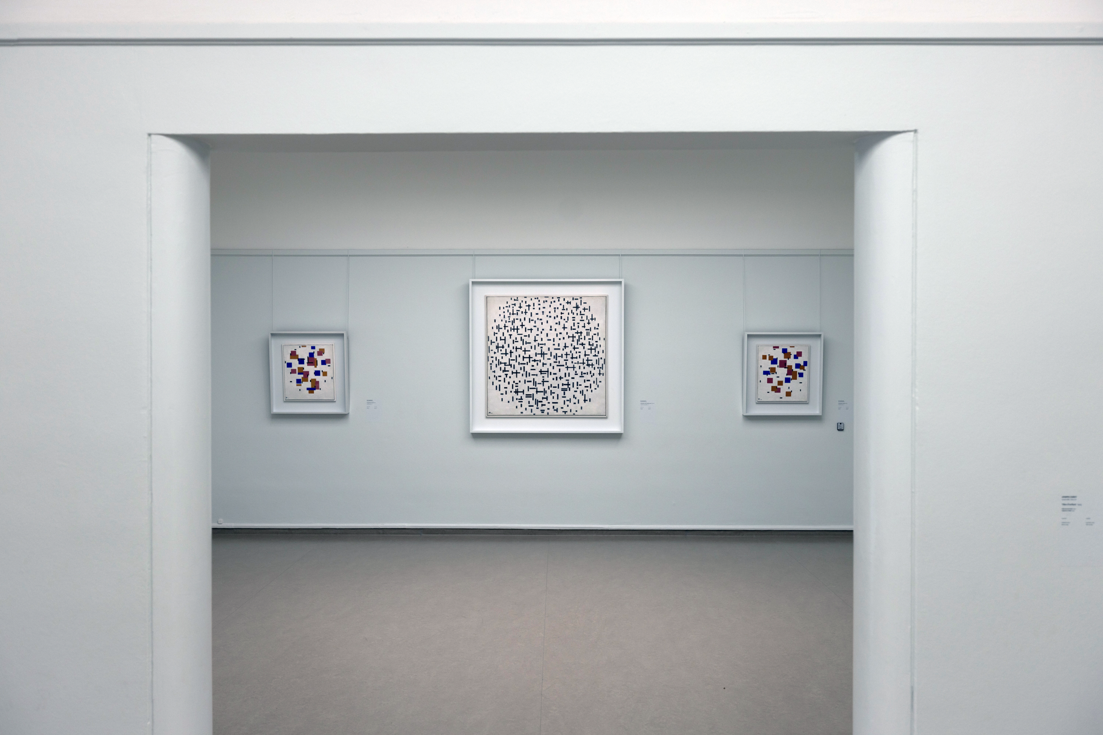 Foto van een zaal in het museum met drie abstracte schilderijen aan een witte muur, waarbij het grootste werk in het midden bestaat uit een patroon van zwarte stippen op een witte achtergrond en de twee kleinere werken aan weerszijden kleurrijke geometrische vormen tonen.