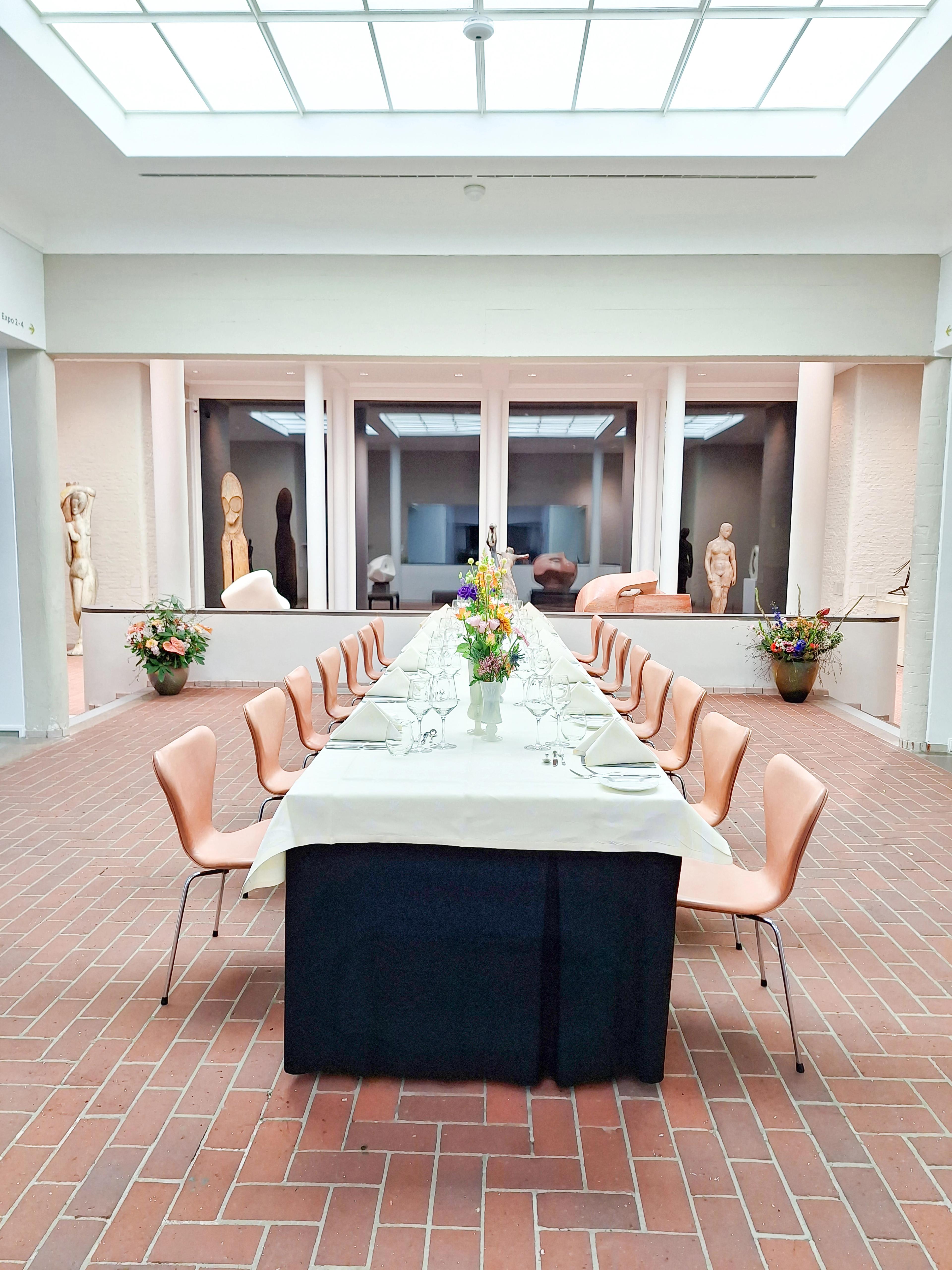 Foto van een gedekte tafel met stoelen in een beeldenzaal van het museum. Op tafel bloemen. Buiten is het donker, het is avond.