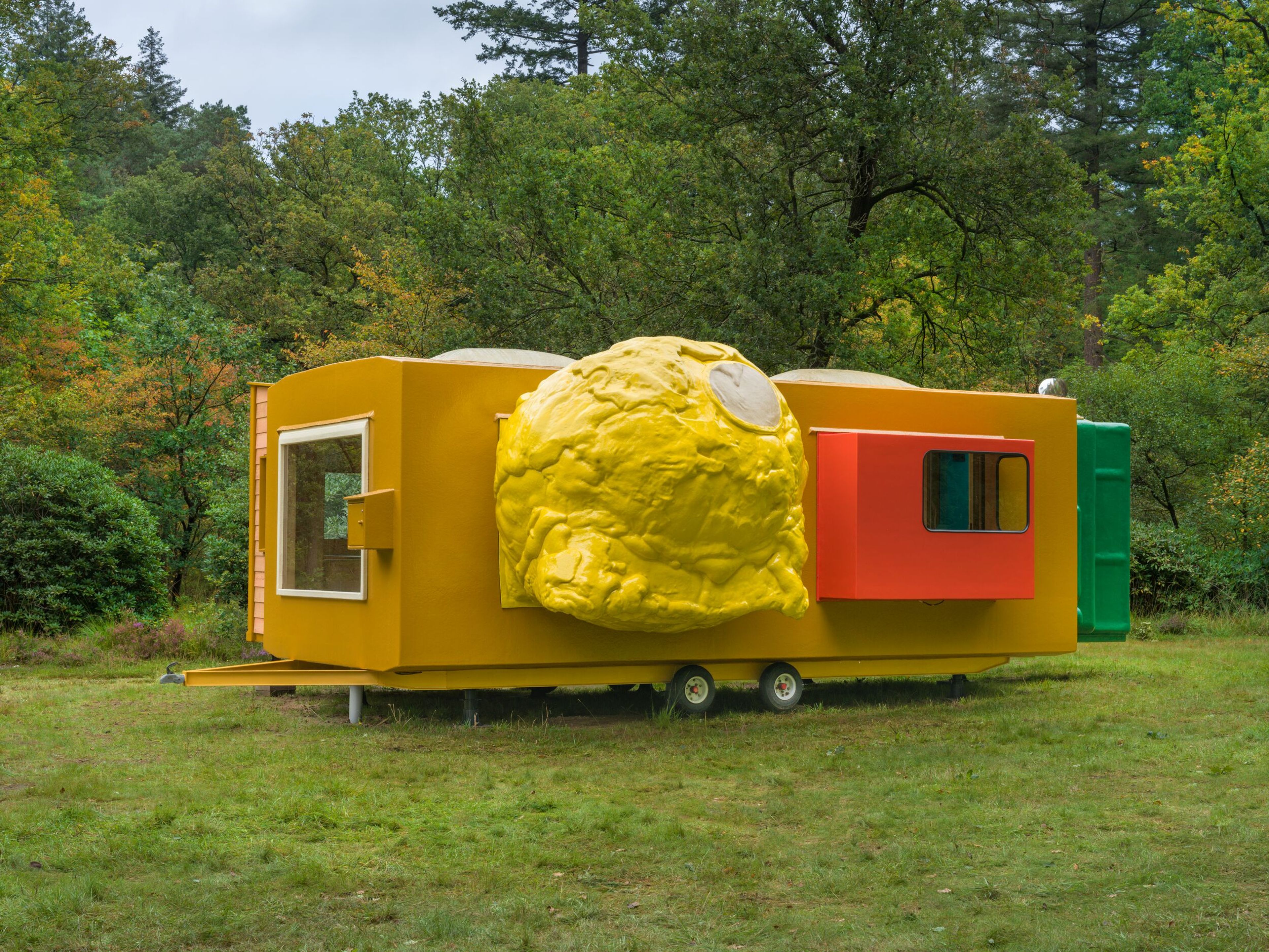 Joep van Lieshout, _Mobile home for Kröller-Müller_, 1995 Foto van een kleurrijk kunstwerk in een bosrijke omgeving met een geel, oranje en groene structuur en een grote gele bolvormige uitbouw aan de zijkant.