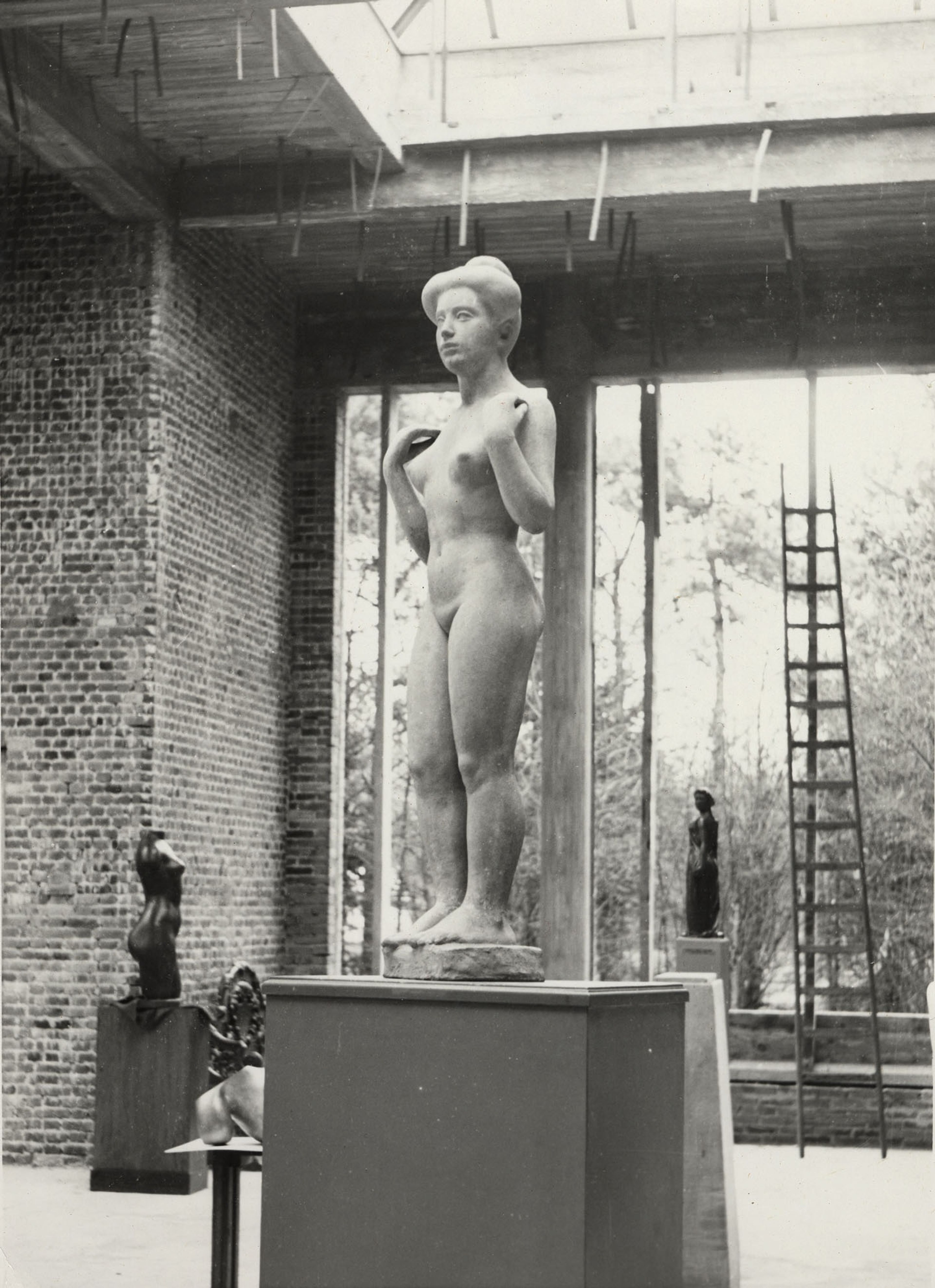 Beeldenzaal, 1953 Zwart-witfoto van de beeldenzaal. Met een naakt vrouwenbeeld, geplaatst op een sokkel in een ruimte met grote ramen en bakstenen muren. Op de achtergrond staan andere beelden en een ladder. De zaal is nog niet af.