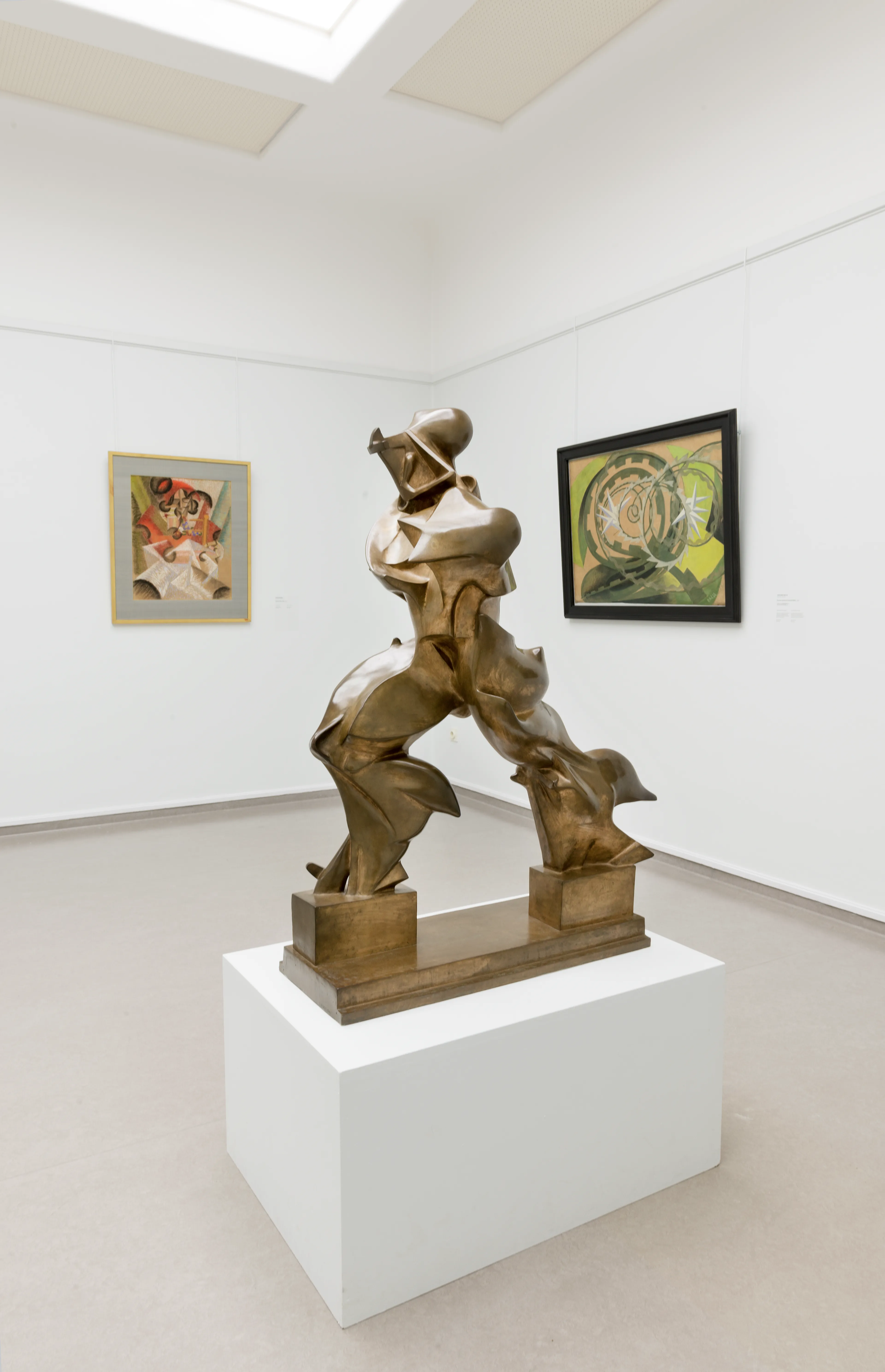 Foto van een tentoonstellingszaal met een bronzen sculptuur centraal geplaatst op een witte sokkel. Twee kleurrijke schilderijen hangen aan witte muren, waarbij sculptuur dynamische, gebogen vormen toont en schilderijen geometrische en organische patronen bevatten.