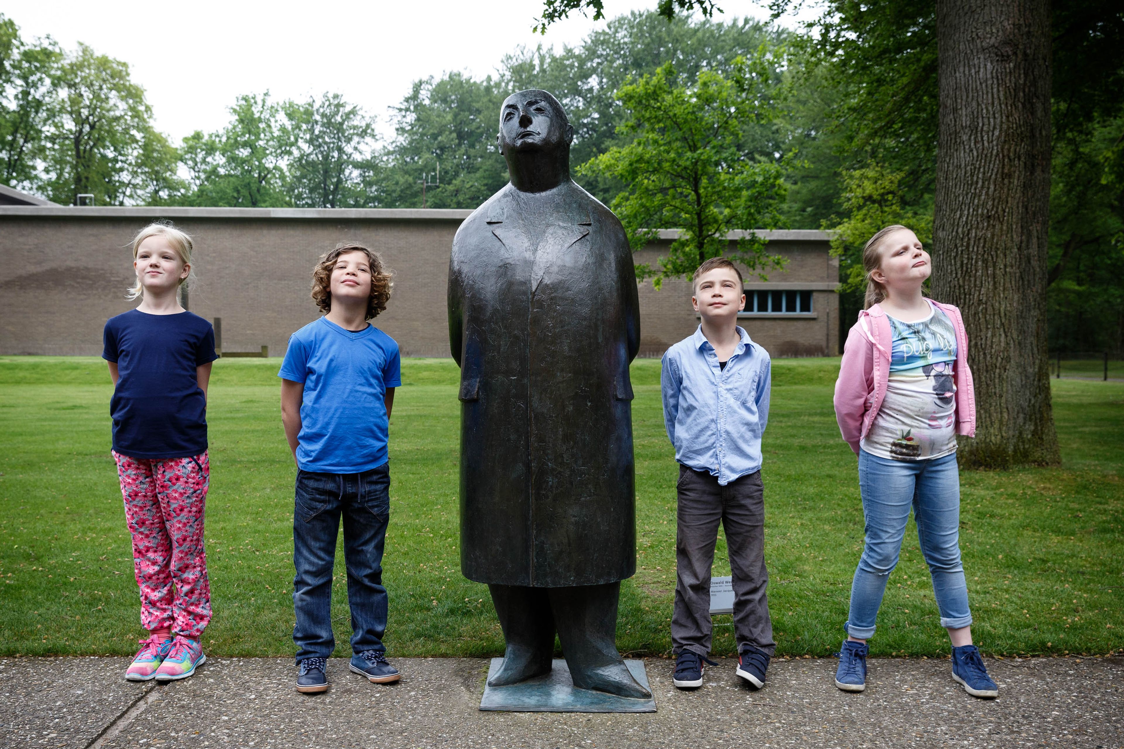 Vier kinderen die naast een bronzen standbeeld van een persoon in een lange jas staan, geplaatst op een betonnen pad in een parkachtige omgeving. Kinderen dragen kleurrijke kleding en staan in een rij met een gebouw en bomen op de achtergrond.