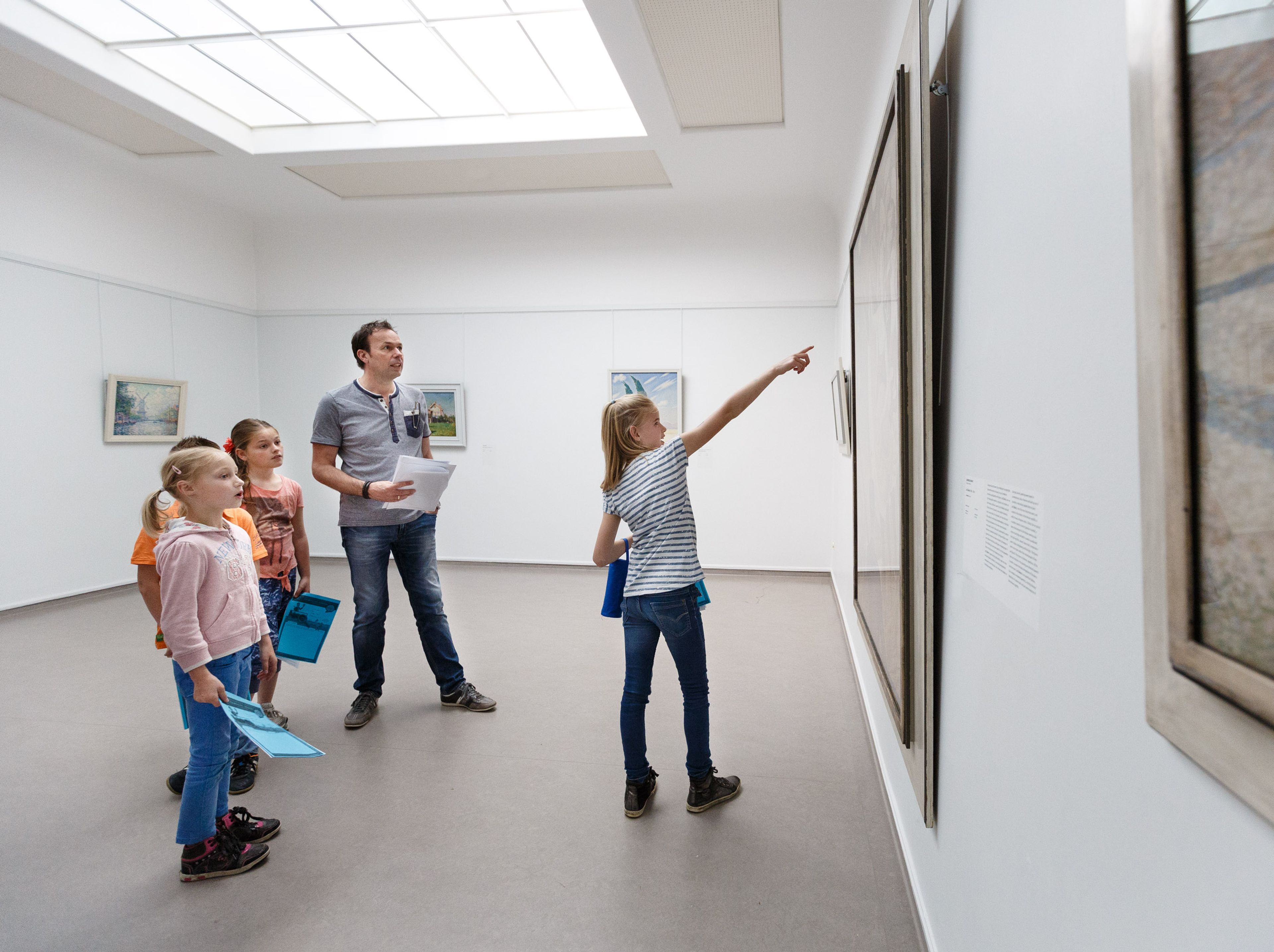 een groep kinderen en een leraar die naar schilderijen aan de muur kijken. Kinderen houden blauwe mappen vast en een meisje wijst naar een kunstwerk, wat een educatieve context en betrokkenheid bij kunst benadrukt.