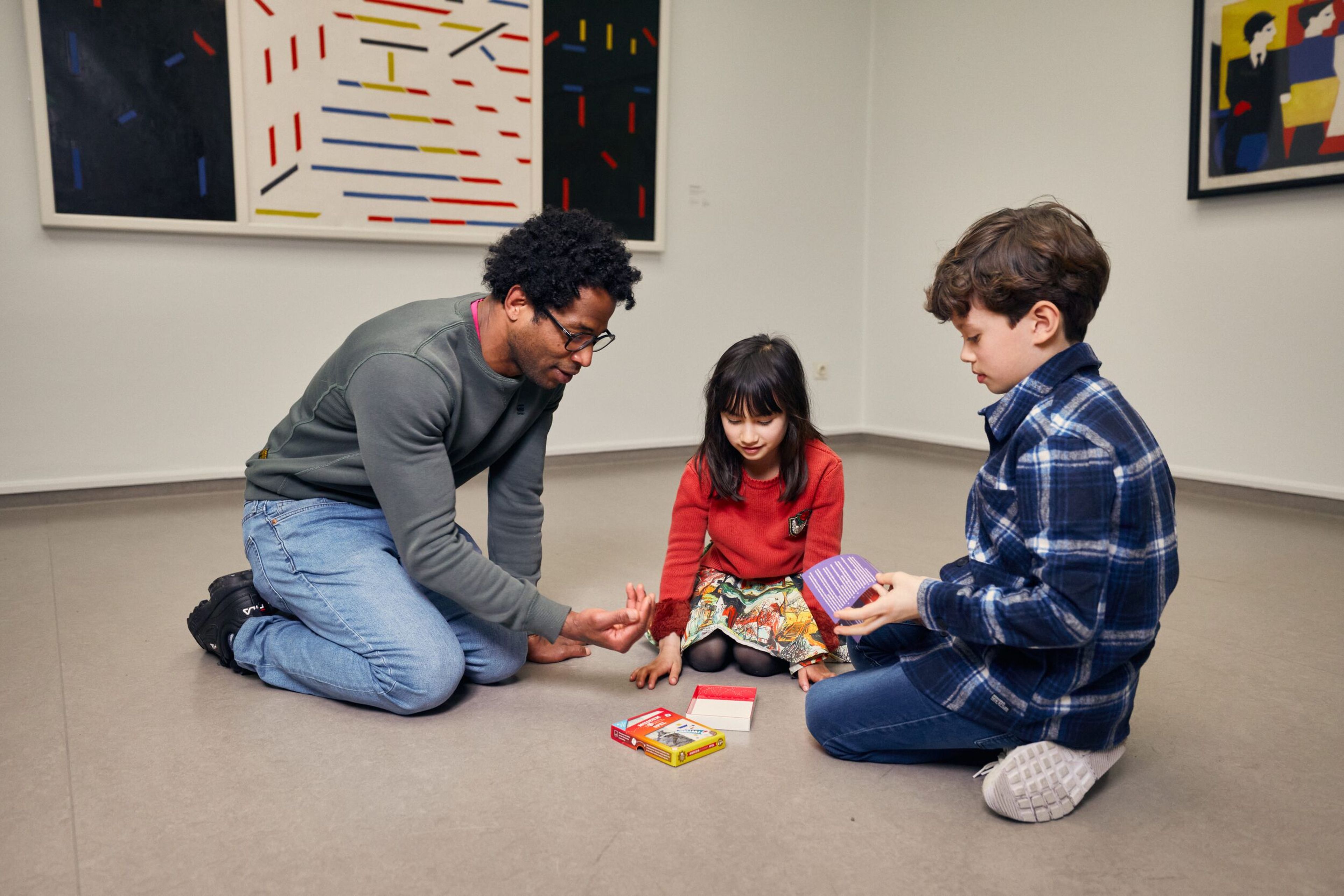 Foto van drie personen die op de grond zitten en samen een spel spelen in een minimalistische kamer met abstracte schilderijen aan de muur. Twee kinderen en een volwassene zijn betrokken bij het spel, waarbij kleurrijke kaarten en een doosje zichtbaar zijn.