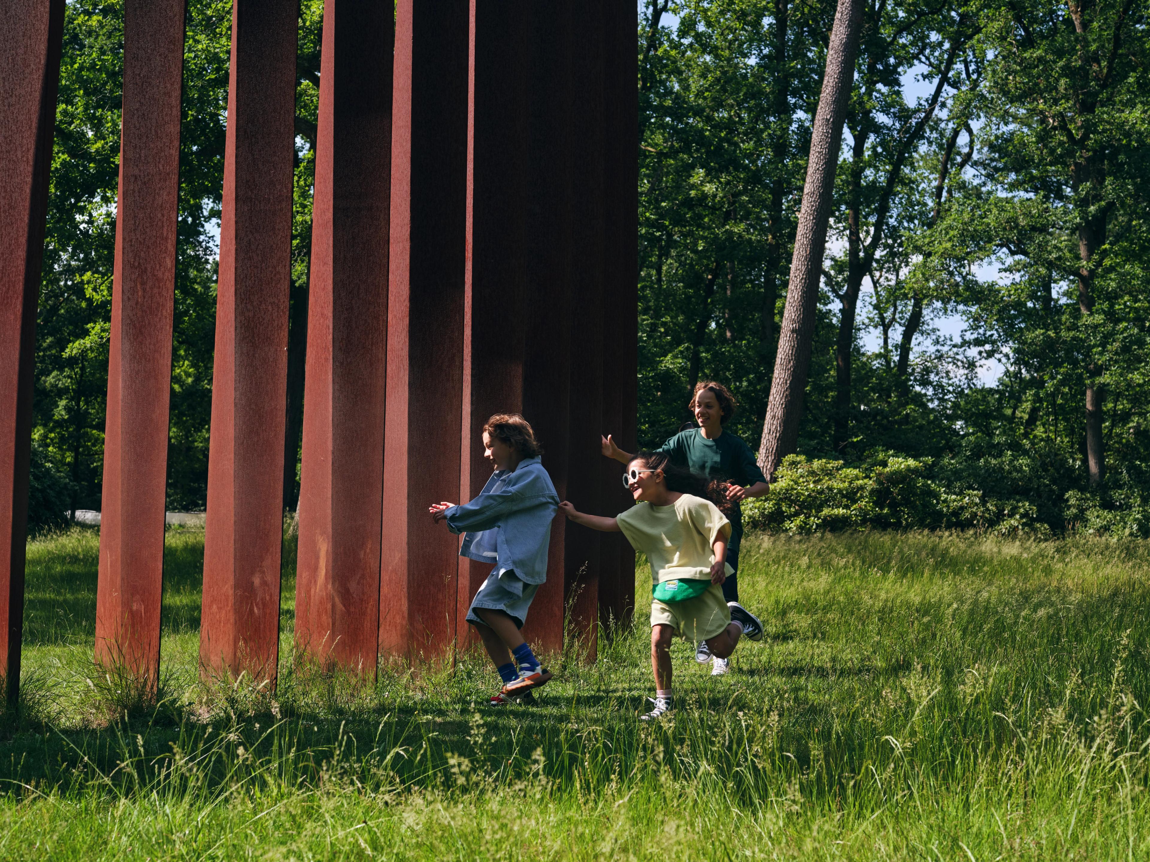Evert Strobos, _Palissade_, 1973-1991 Foto van drie kinderen die rennen en spelen bij hoge, roestkleurige metalen pilaren in een bosrijke omgeving met gras