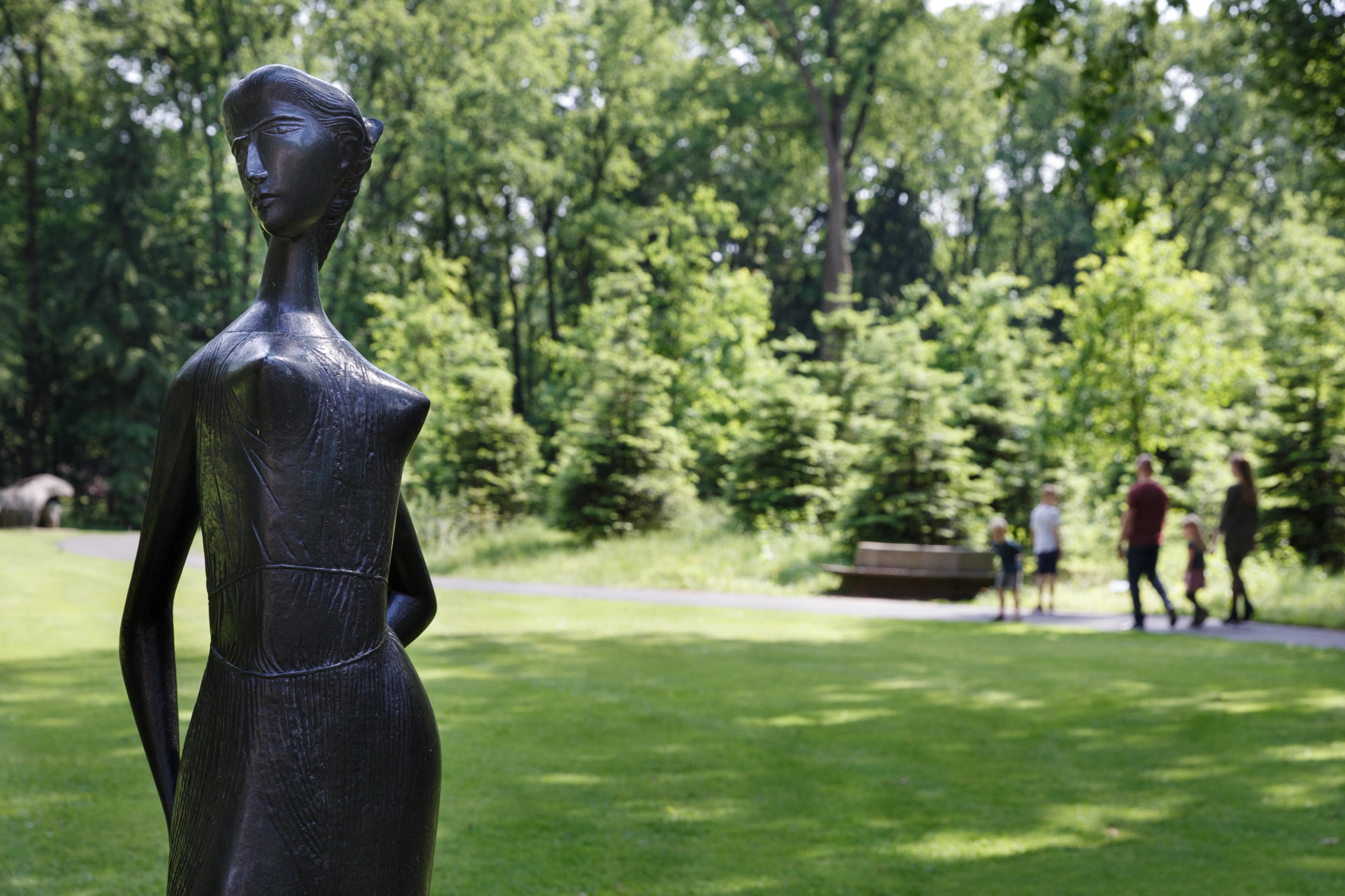 Foto van een abstracte, donkere bronzen beeld van een vrouw in de beeldentuin. Met groen gras en bomen op de achtergrond. Op een pad lopen vier mensen.