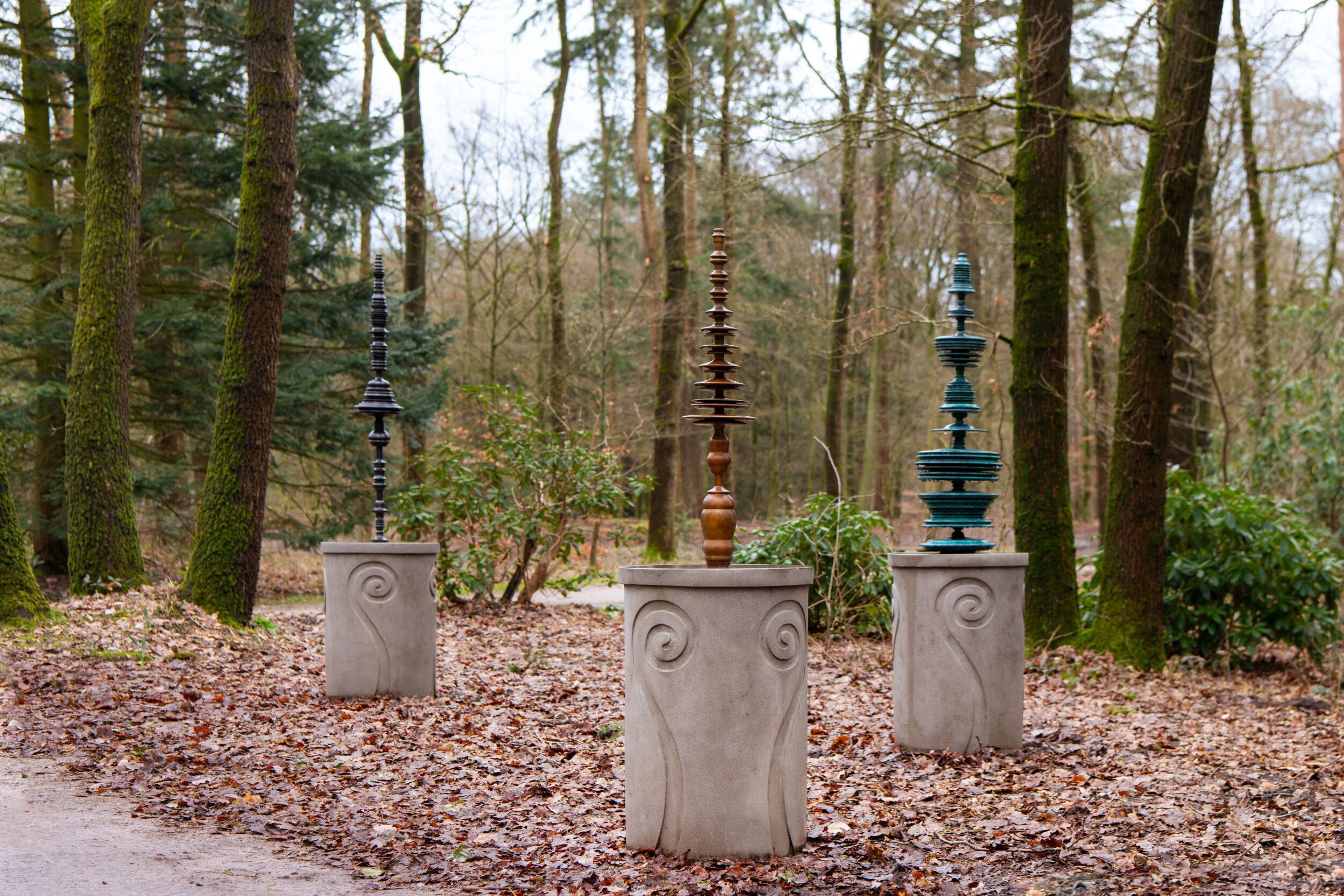 A photograph of three sculptures in a wooded setting, each consisting of a concrete plinth topped by a vertical stack of different shapes and colours.