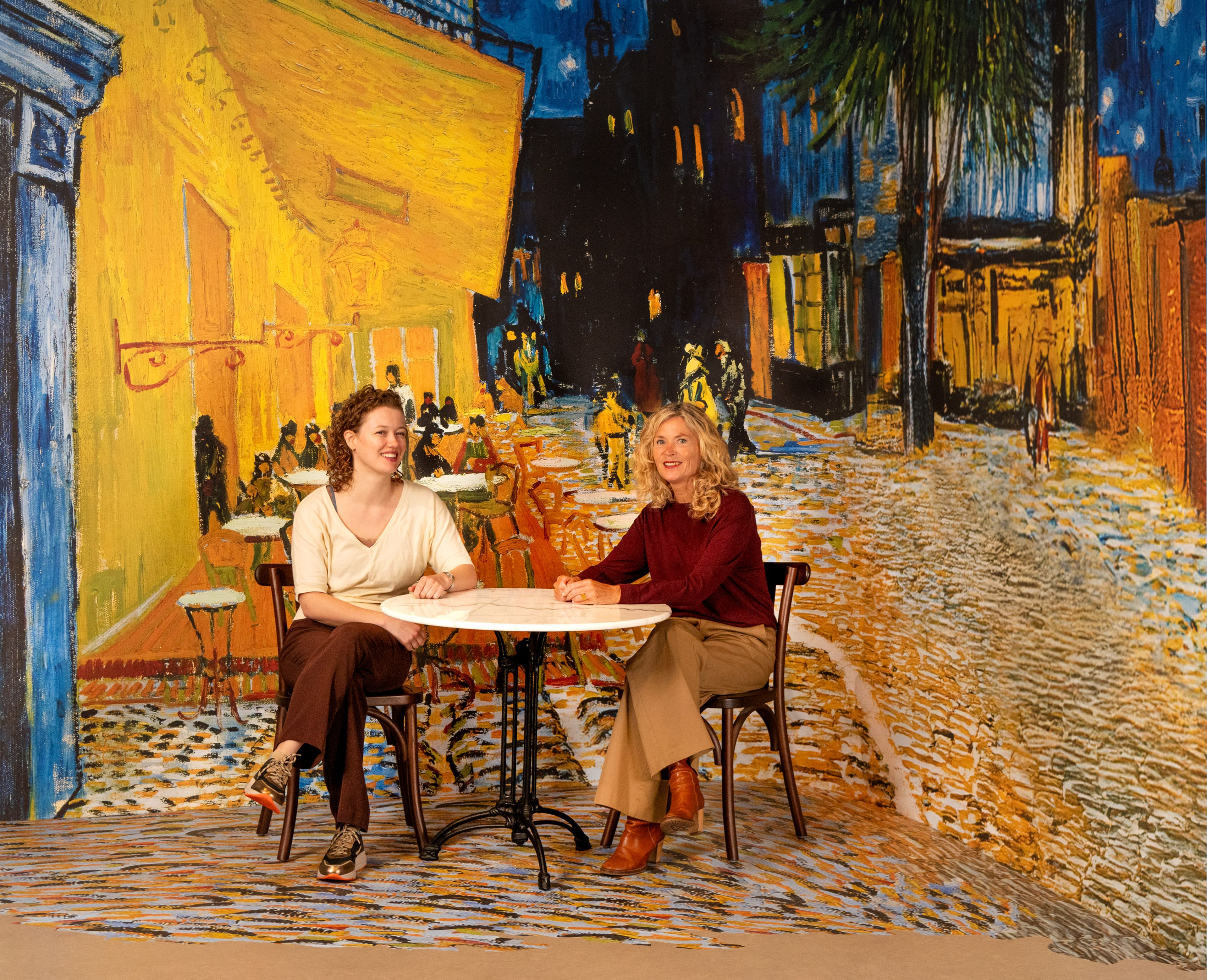 Twee bezoekers zitten op een terrasstoel, voor een afbeelding van Van Goghs ‘Caféterras bij nacht’