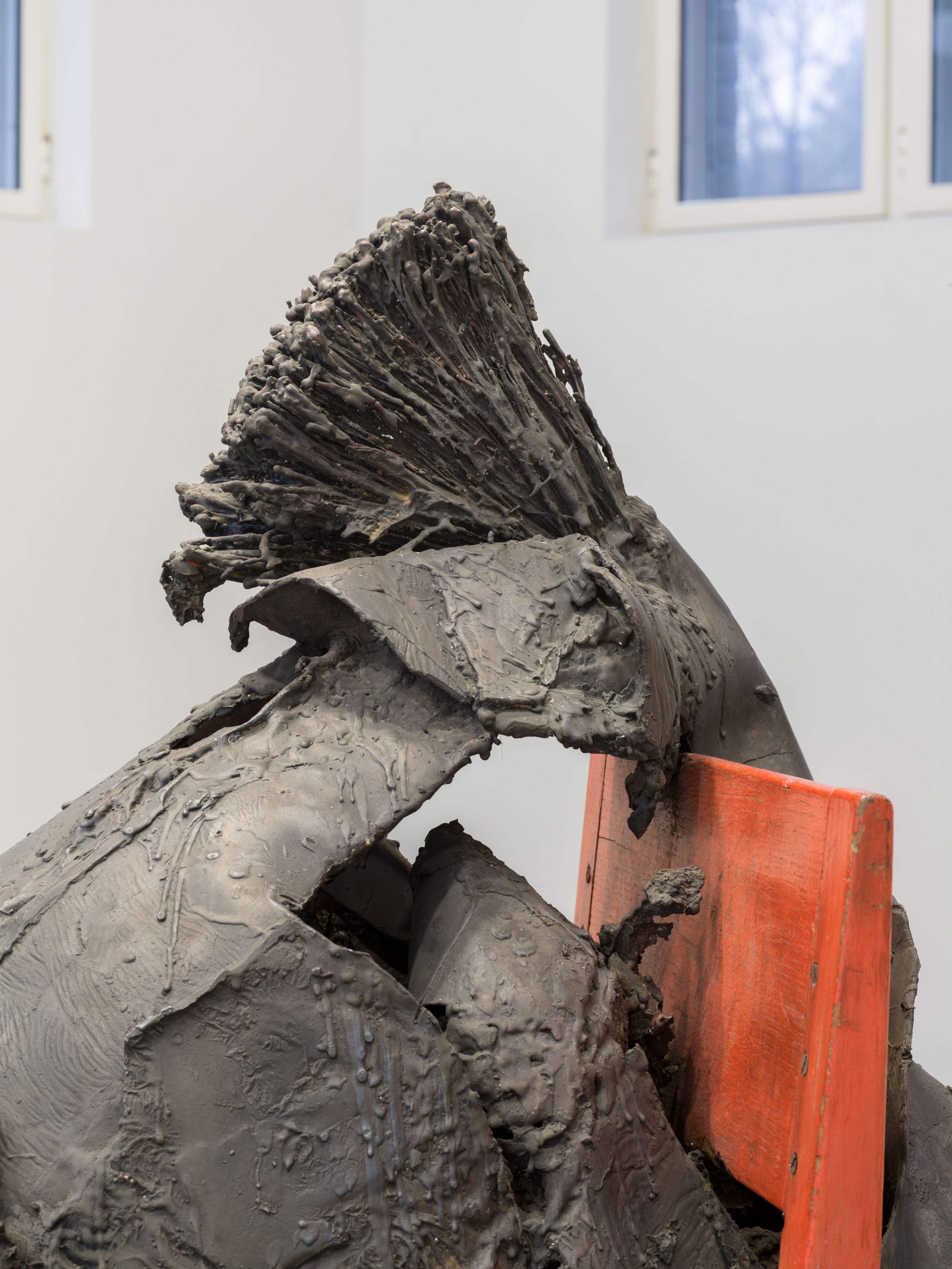 Foto van een abstracte sculptuur met ruwe, textuurrijke oppervlakken en een opvallend, waaierachtig element bovenaan. Sculptuur staat op een oranje houten stoel in een lichte kamer met ramen op de achtergrond.