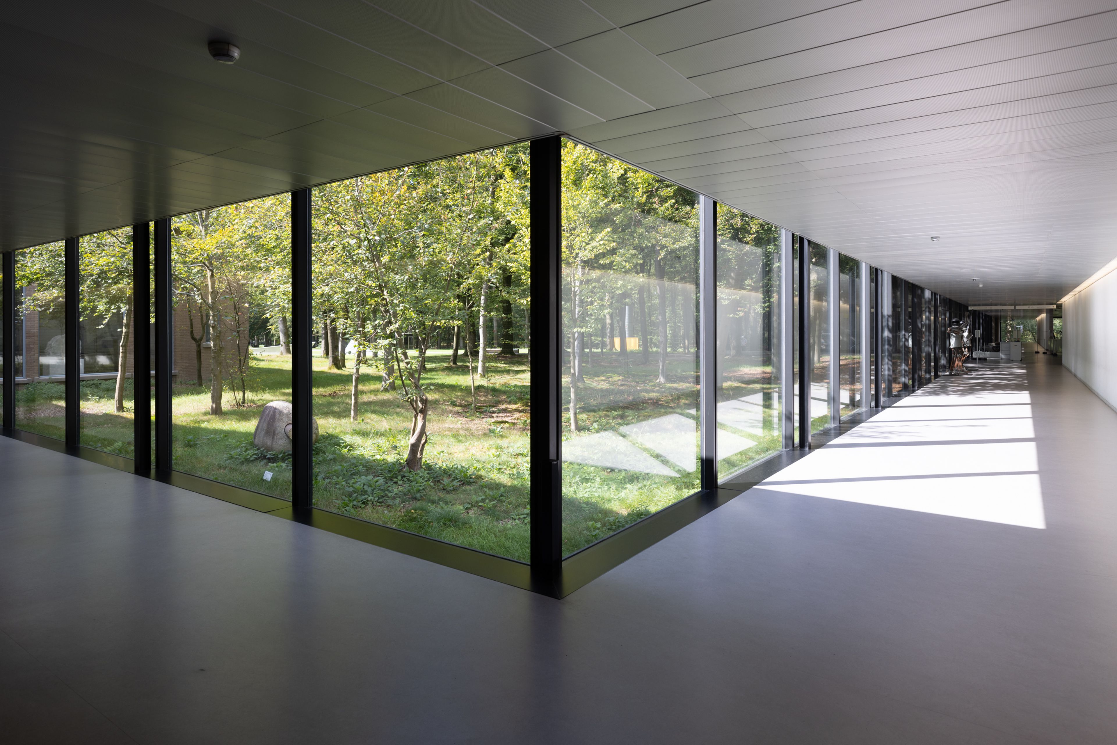 Gang met glazen wanden in het Kröller-Müller Museum Fotografie van een modern gebouw met grote glazen wanden die uitzicht bieden op een groene tuin met bomen en gras. Zonlicht valt door ramen en creëert reflecties en schaduwen op de vloer