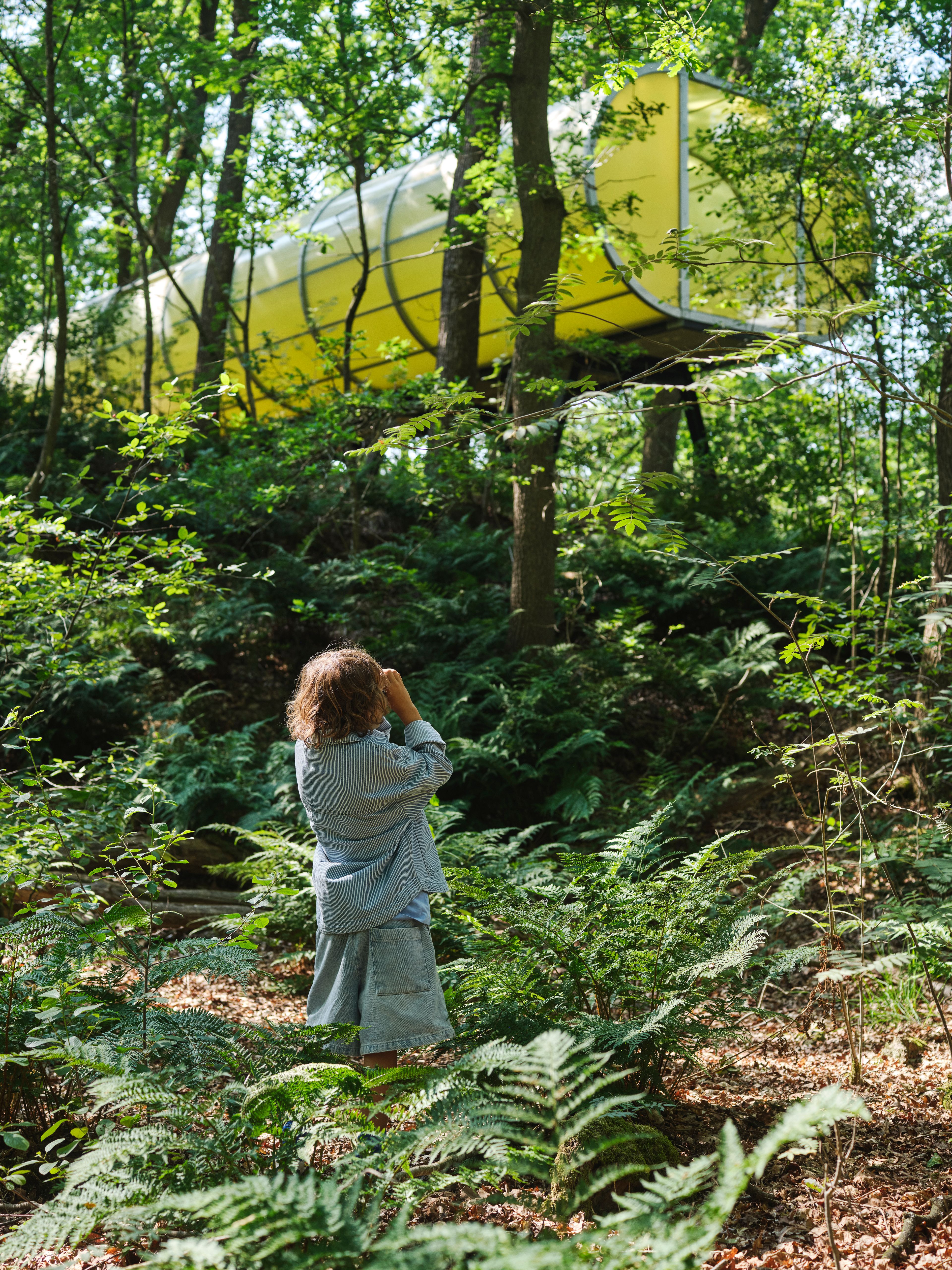 Foto waarop een kind in een dicht bos staat en een foto maakt van een geel kunstwerk tussen de bomen.