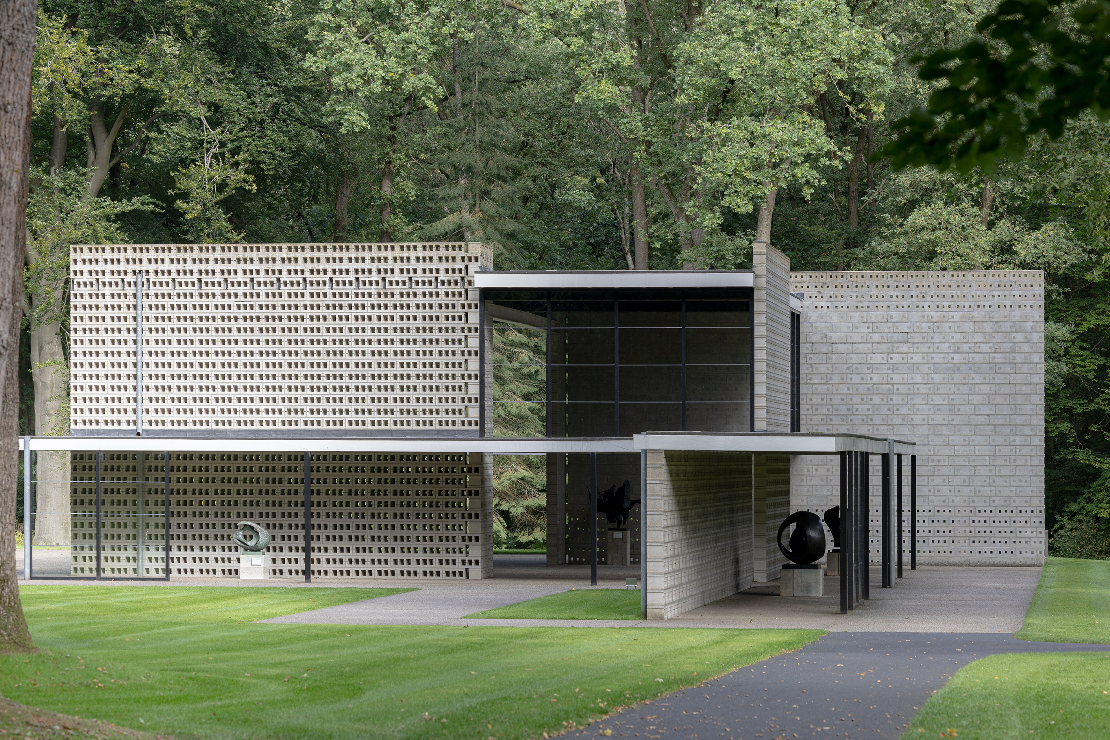 Gerrit Rietveld, _Rietveldpaviljoen_, 1954-1955 (herbouwd in 1964-1965 en 2010) Foto van het Rietveldpaviljoen. Een modern gebouw met strakke lijnen en witte bakstenen, in de groene parkachtige omgeving van de beeldentuin, met bomen op de achtergrond. Voor het gebouw staat een abstract kunstwerk op een sokkel.