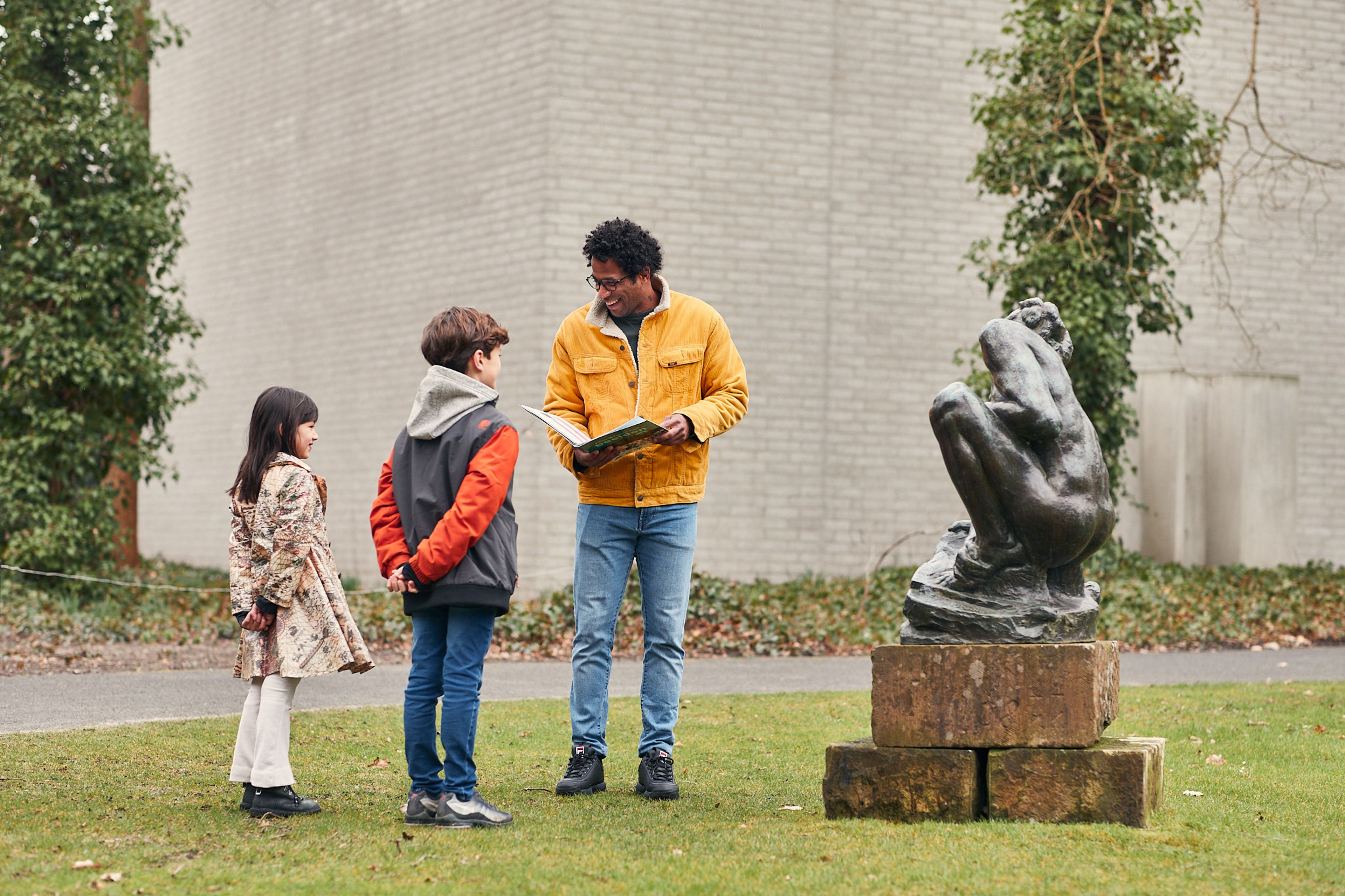 Drie personen staan buiten bij een bronzen beeld op een sokkel, met een man die een boek vasthoudt en twee kinderen die naar het beeld kijken. De setting toont een educatieve of informele uitlegmoment in een tuin of park met een bakstenen muur en groen rondom.