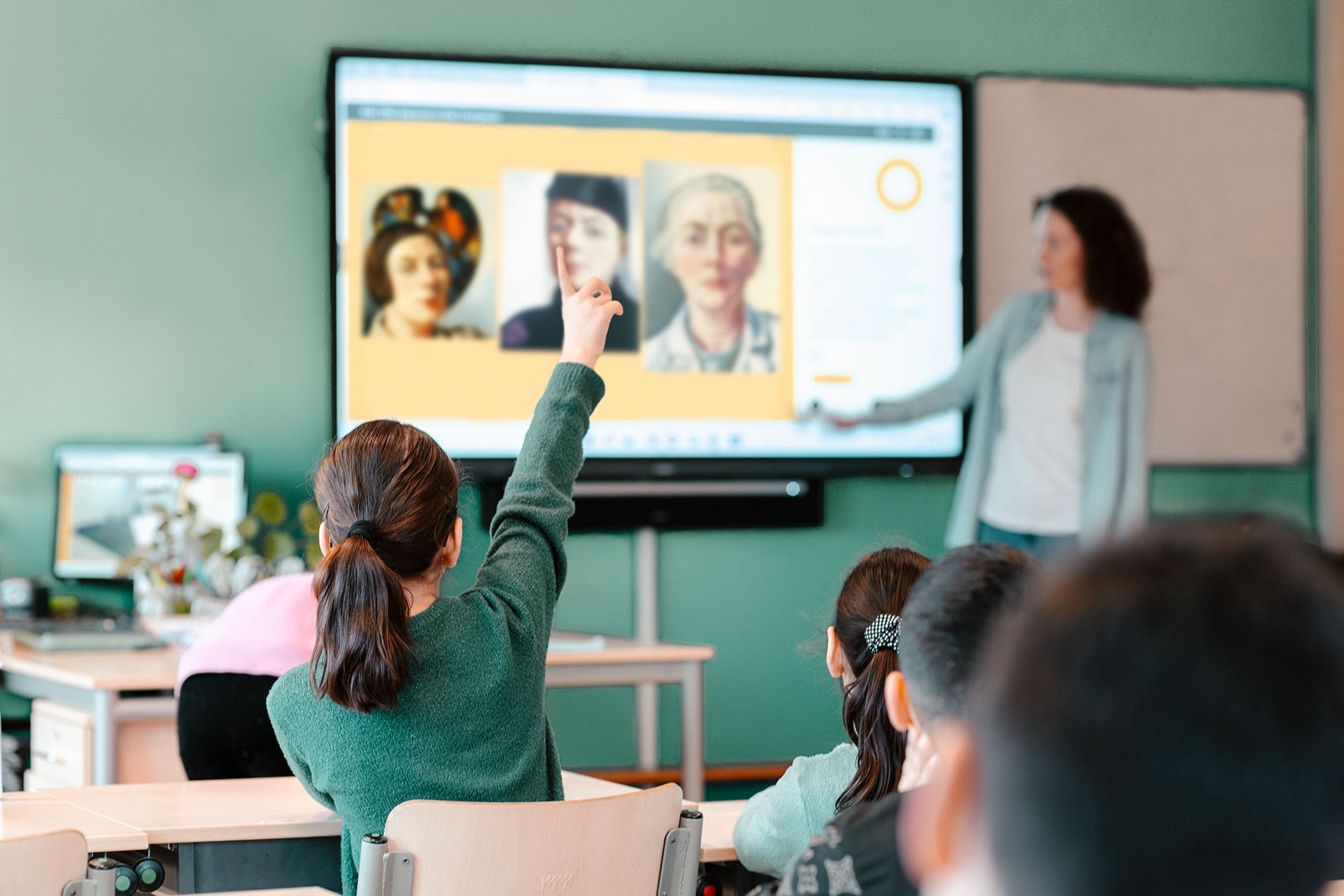 Foto van klaslokaal met leerlingen die naar digitaal scherm kijken waar gezichten van mensen worden getoond. Docent wijst naar het scherm. Leerling op de voorgrond steekt hand op, wat interactie en betrokkenheid benadrukt.