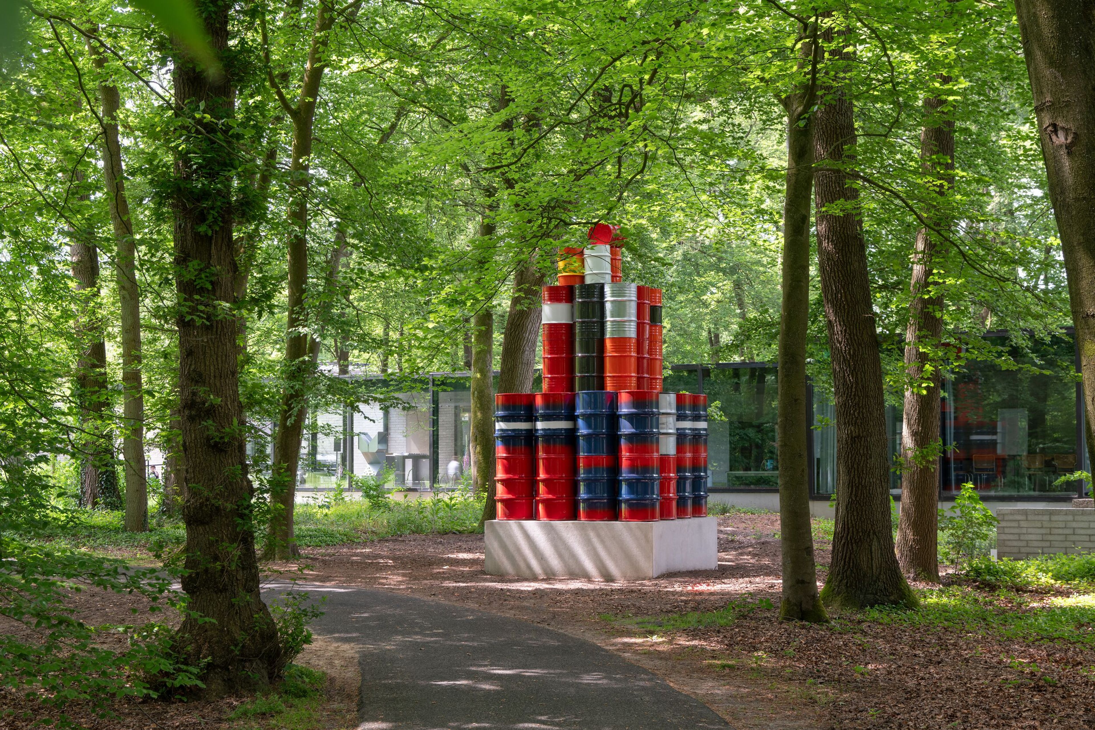 kunstwerk bestaande uit olievaten in de kleuren rood, blauw en wit. Op een witte sokkel, in een bosrijke omgeving met groen blad aan de bomen. Op de achtergrond een modern museumgebouw