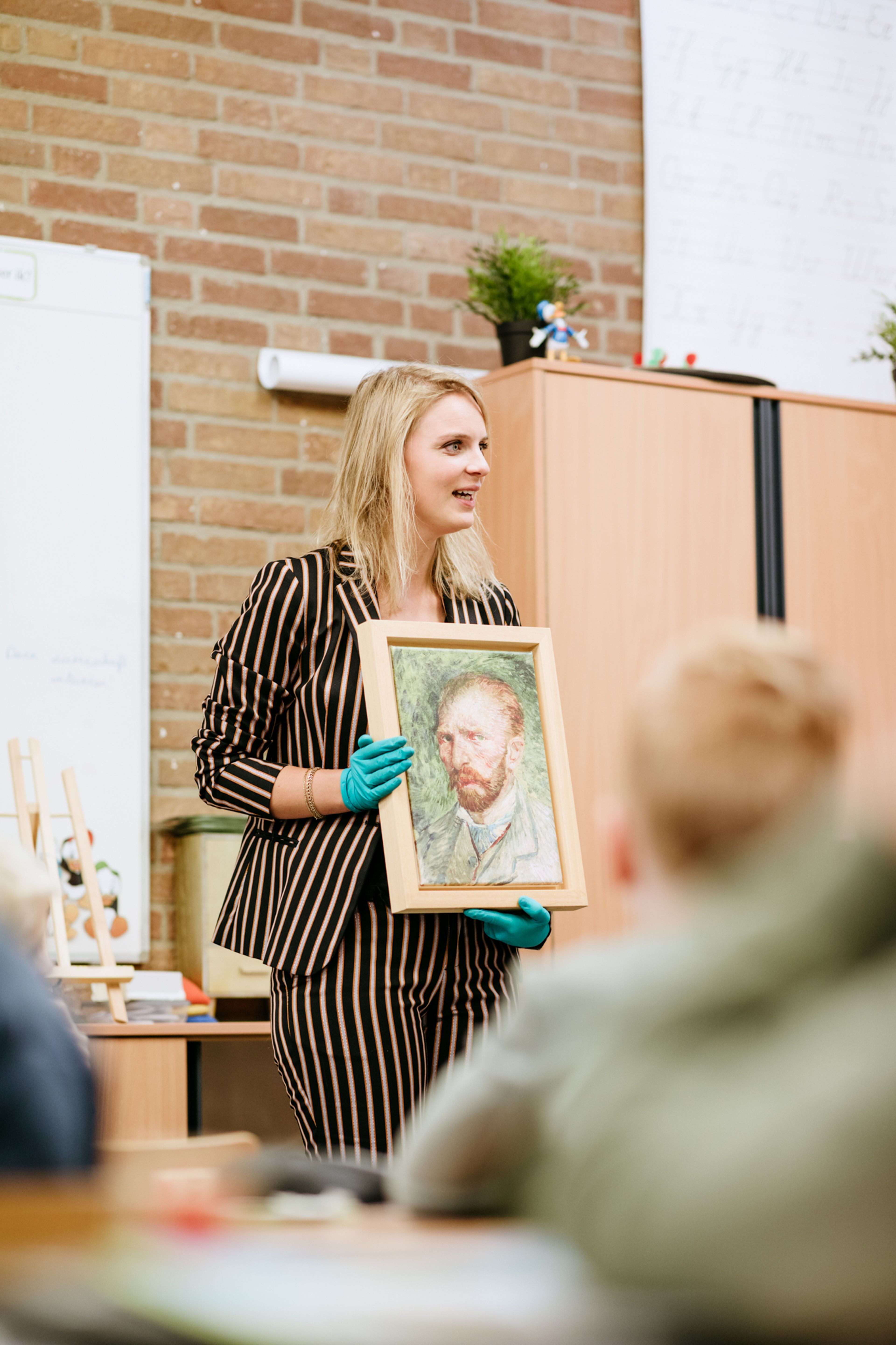 Museumdocent houdt een reproductie van 'Zelfportret' van Vincent van Gogh vast. Op de voorgrond leerlingen in de klas. 