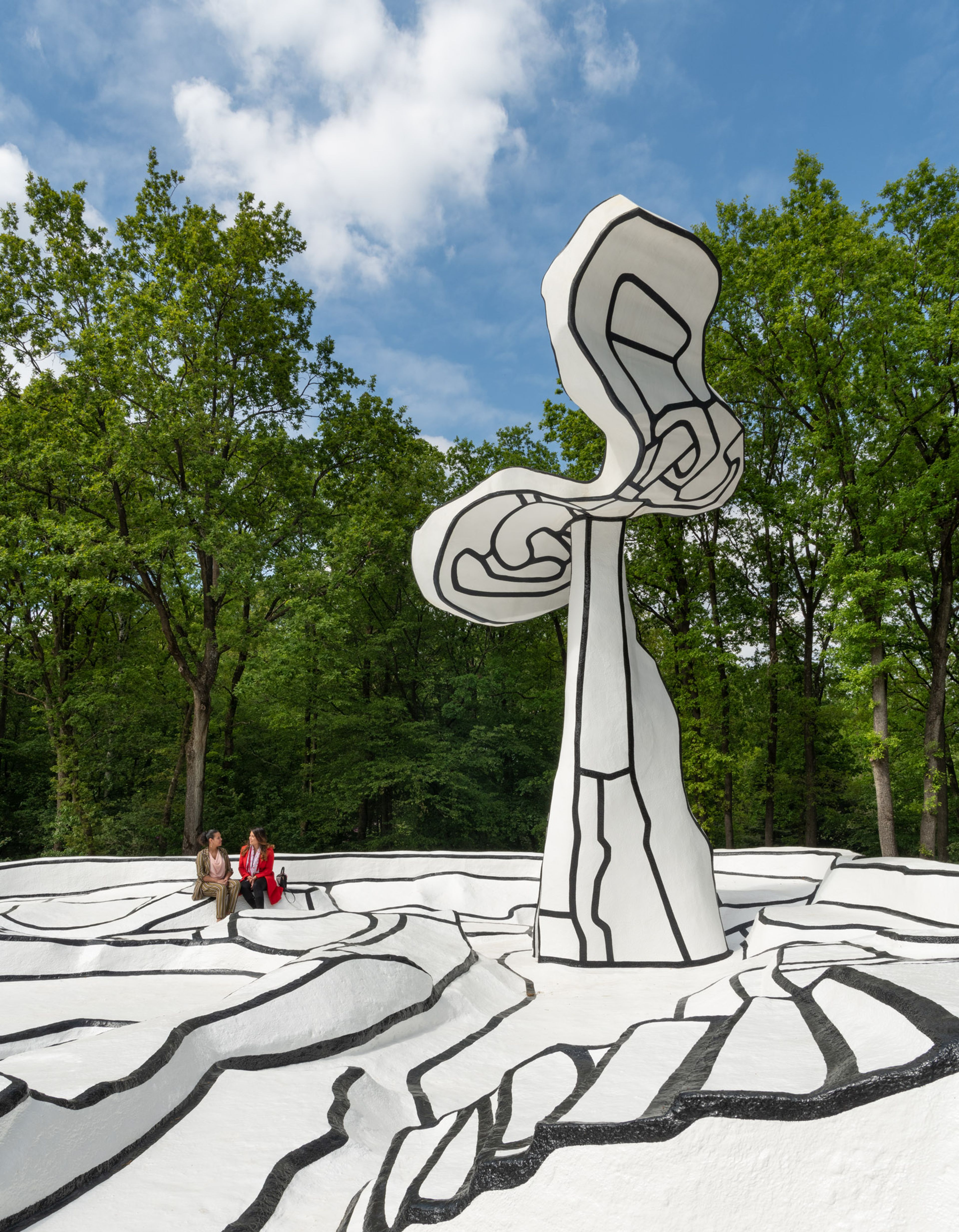 Foto van een buitensculptuur met abstracte, zwart-witte lijnen die een complex patroon op de grond vormen, omgeven door groene bomen en blauwe lucht. Drie mensen zitten op de sculptuur
