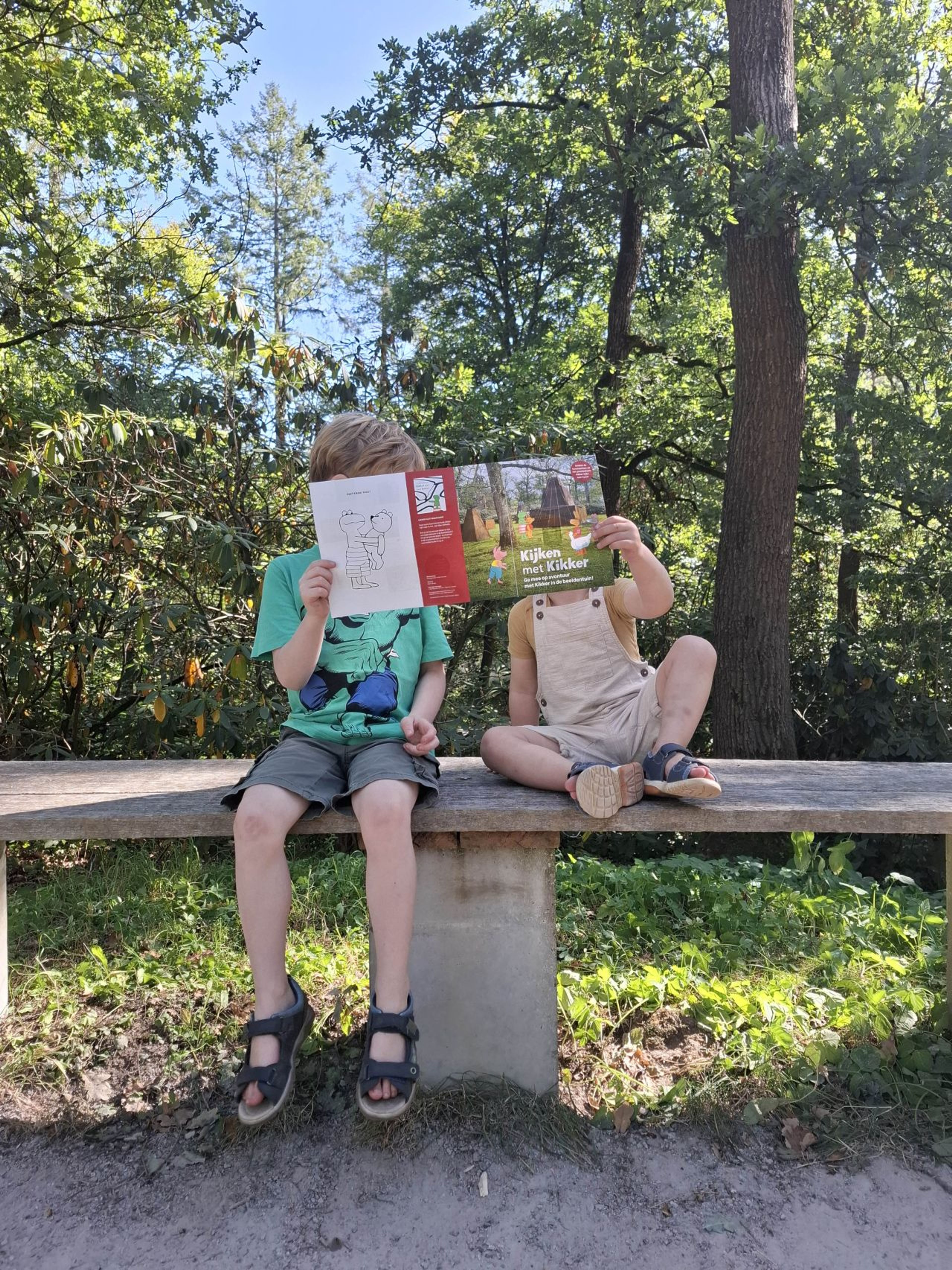 Foto van twee kinderen die op een stenen bank in een bosrijke omgeving zitten en samen een folder voor hun gezicht houden. Op de folder staat ‘Kijken met Kikker’.