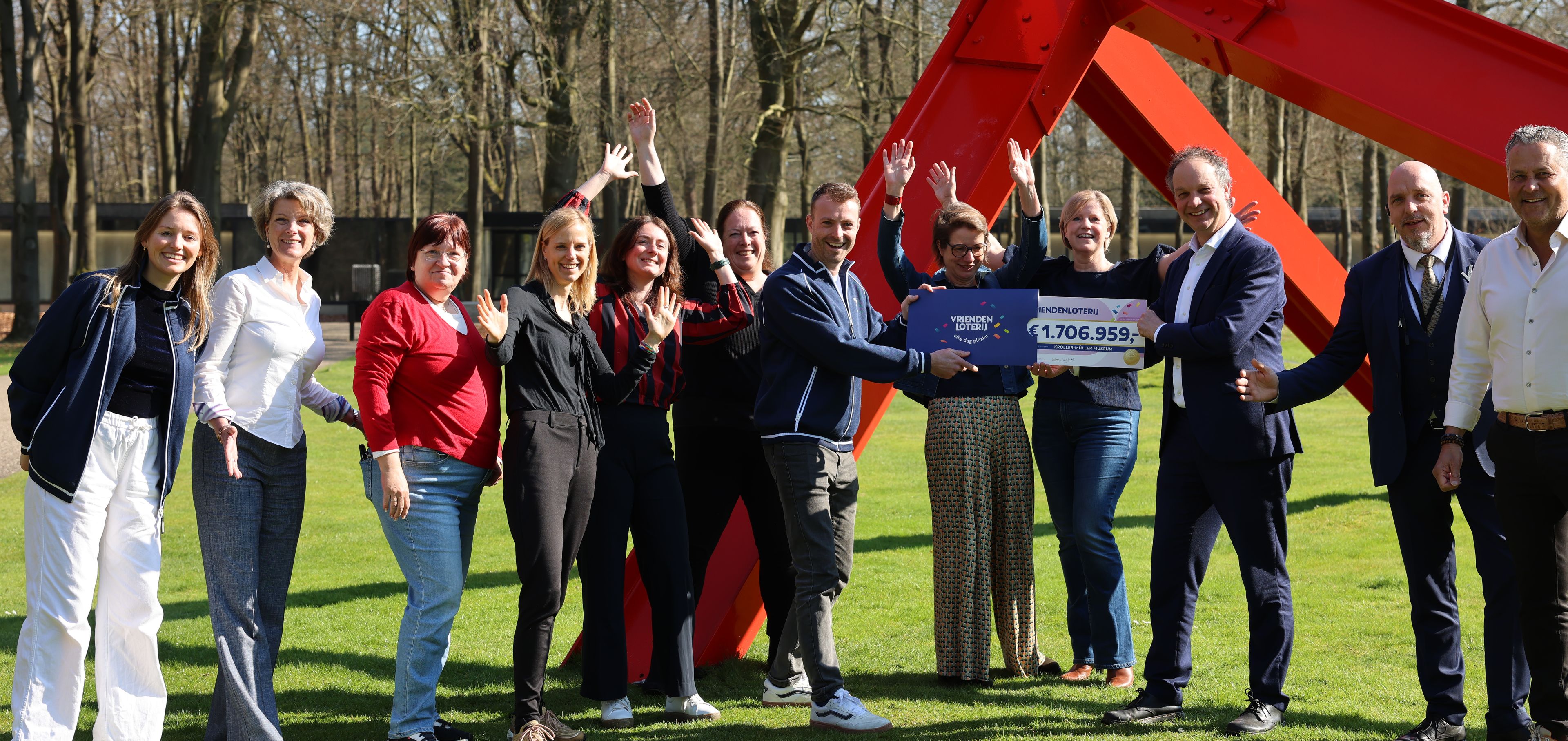 Medewerkers van het museum ontvangen een cheque van de VriendenLoterij, ze staan vrolijk bij een rood kunstwerk in de buitenlucht