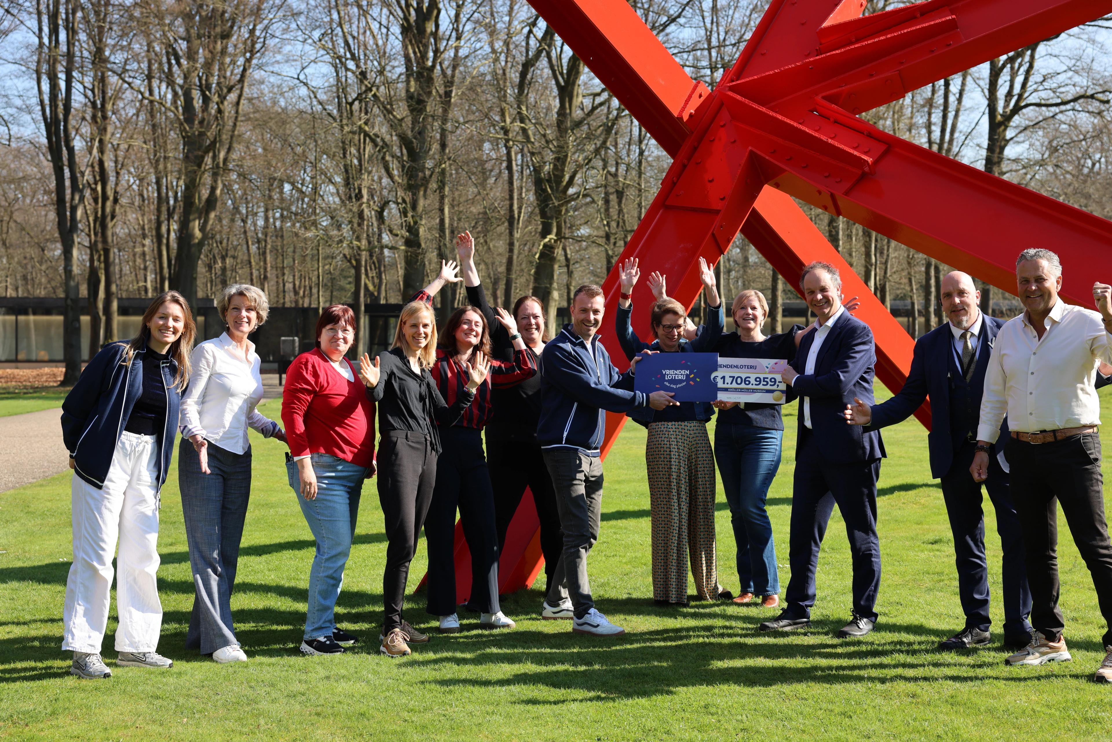 Medewerkers van het museum ontvangen een cheque van de VriendenLoterij, ze staan vrolijk bij een rood kunstwerk in de buitenlucht