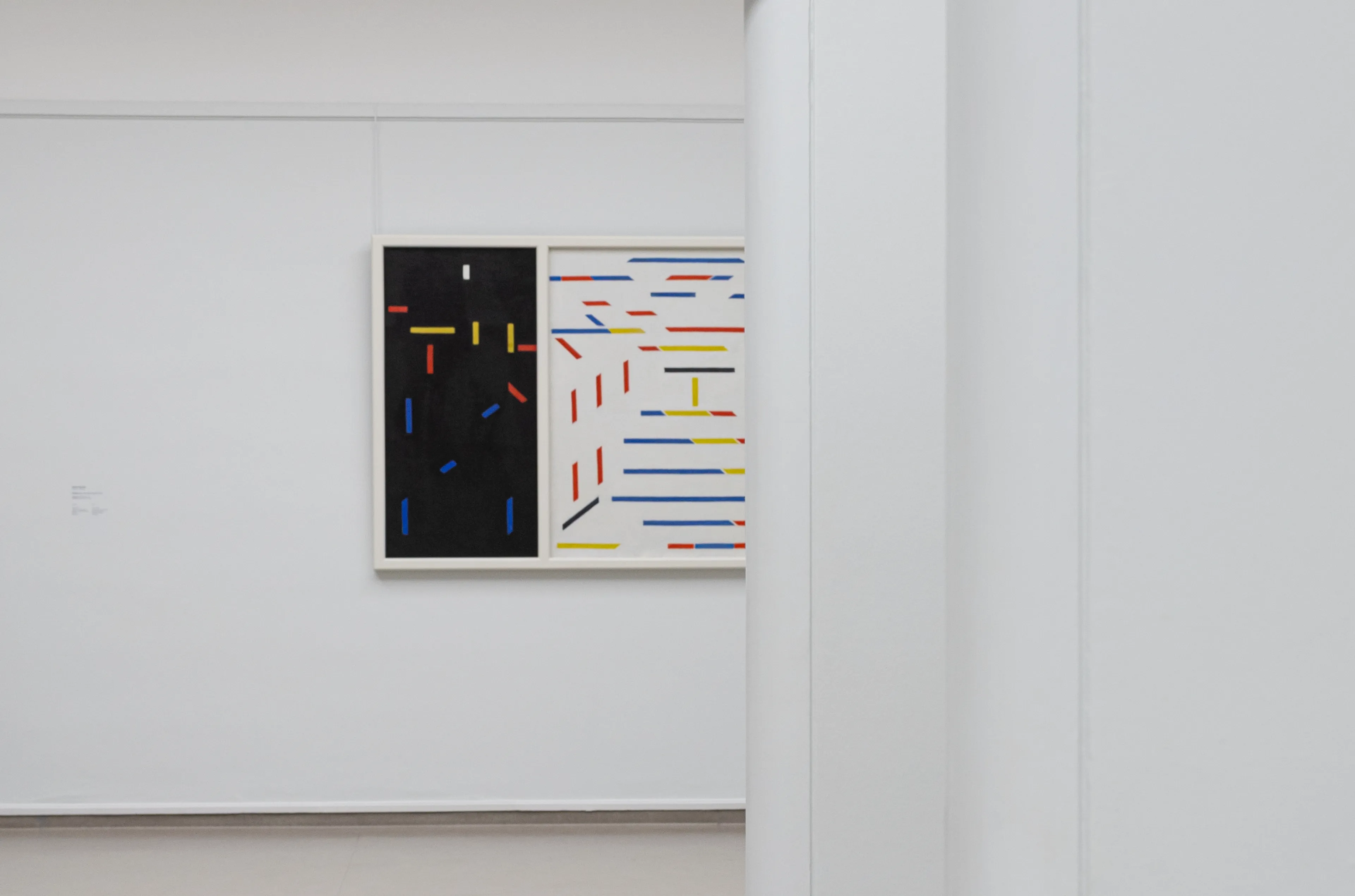 Fotografie van een museuminterieur met witte muren en een abstract schilderij bestaande uit gekleurde lijnen op een zwart en wit vlak.