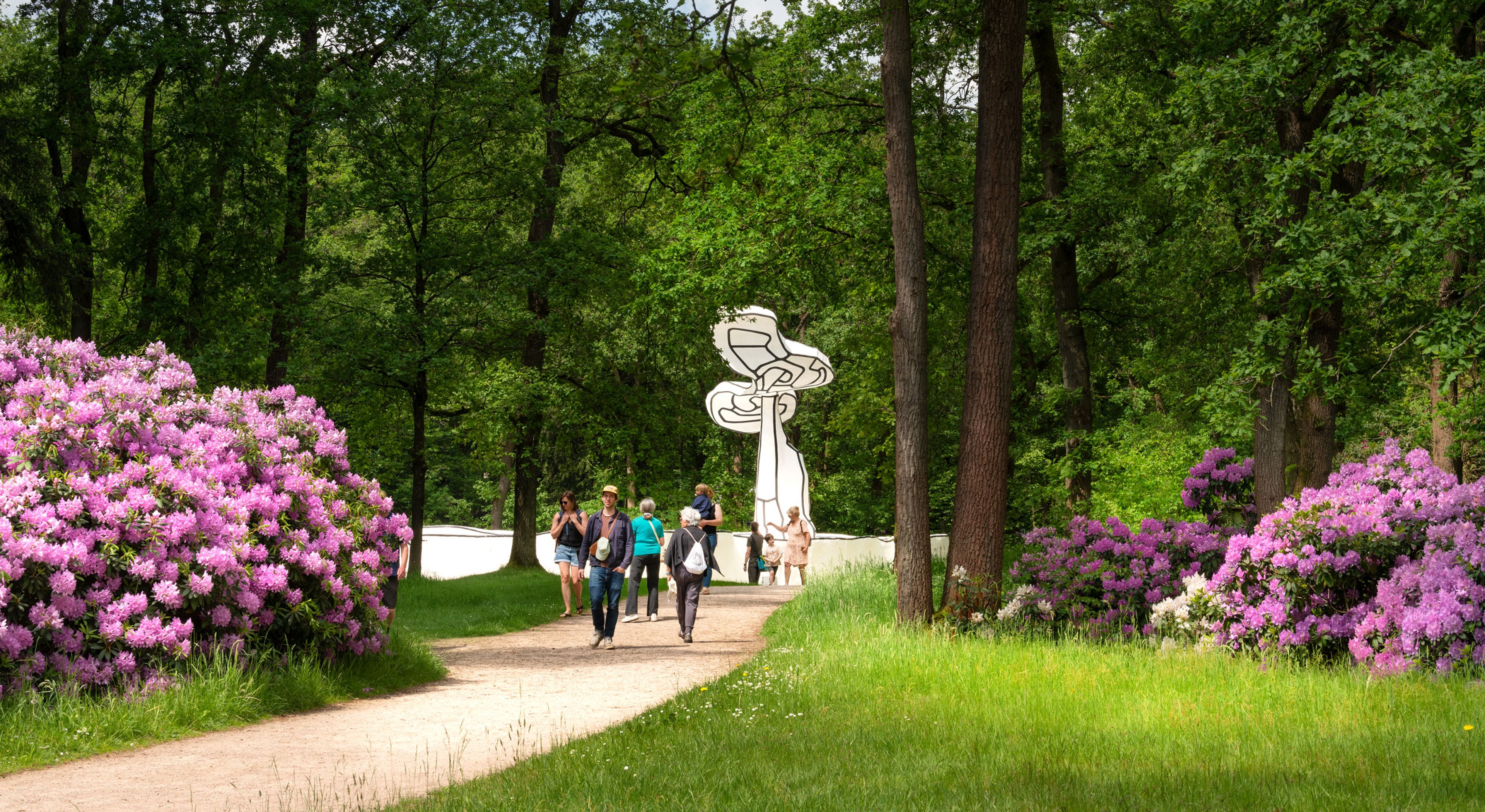Bezoekers lopen over een pad in de beeldentuin. Op de achtergrond een zwart en wit kunstwerk. Naast het pad bloeien rododendronstruiken