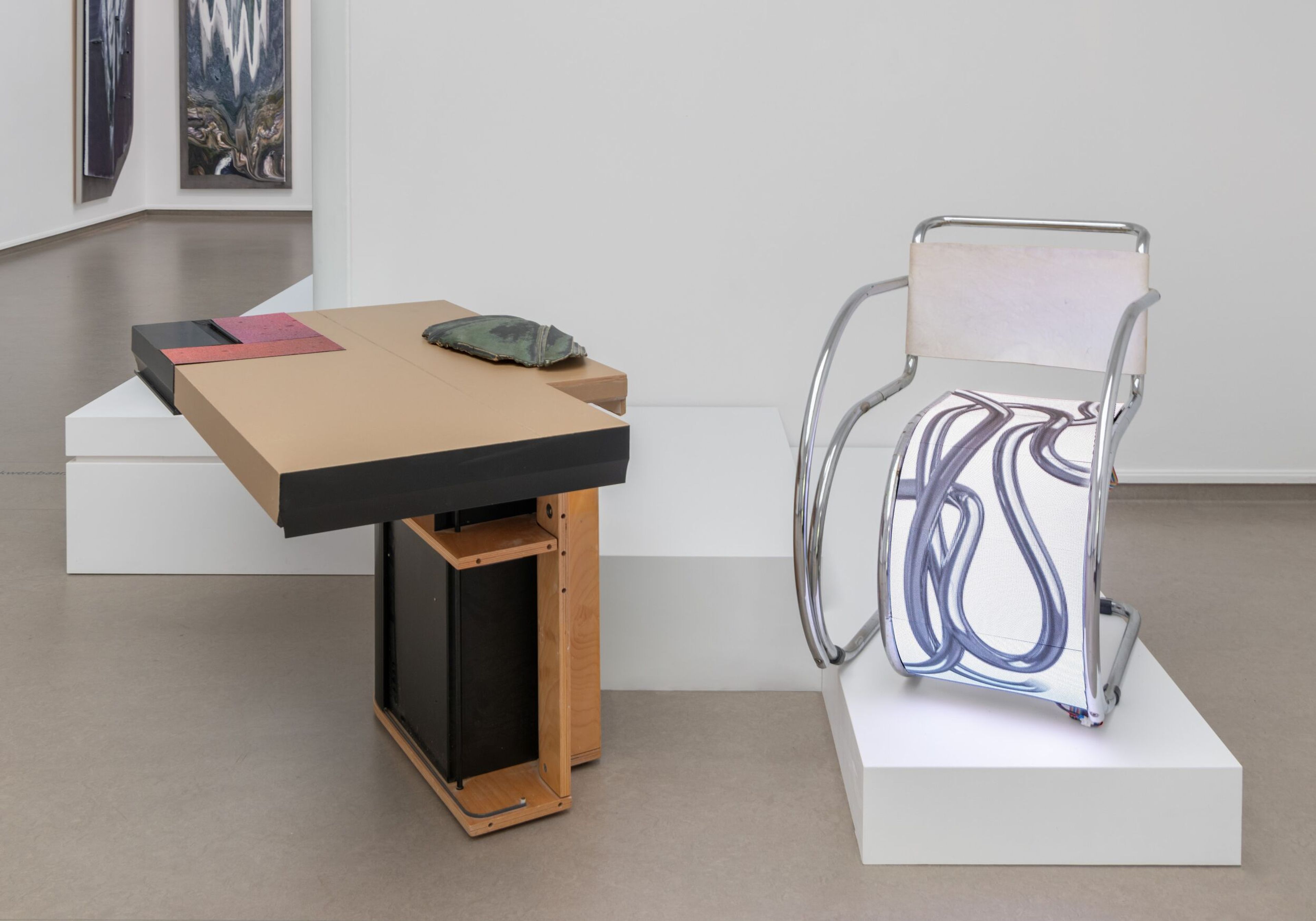 Lilian Kreutzberger. RAUFASER, 2025 Foto van een tentoonstelling met twee sculpturen op witte sokkels. Linkerobject bestaat uit een houten constructie met een groen voorwerp erop, rechterobject is een metalen stoel met een transparante zitting en rugleuning met abstracte blauwe lijnen.