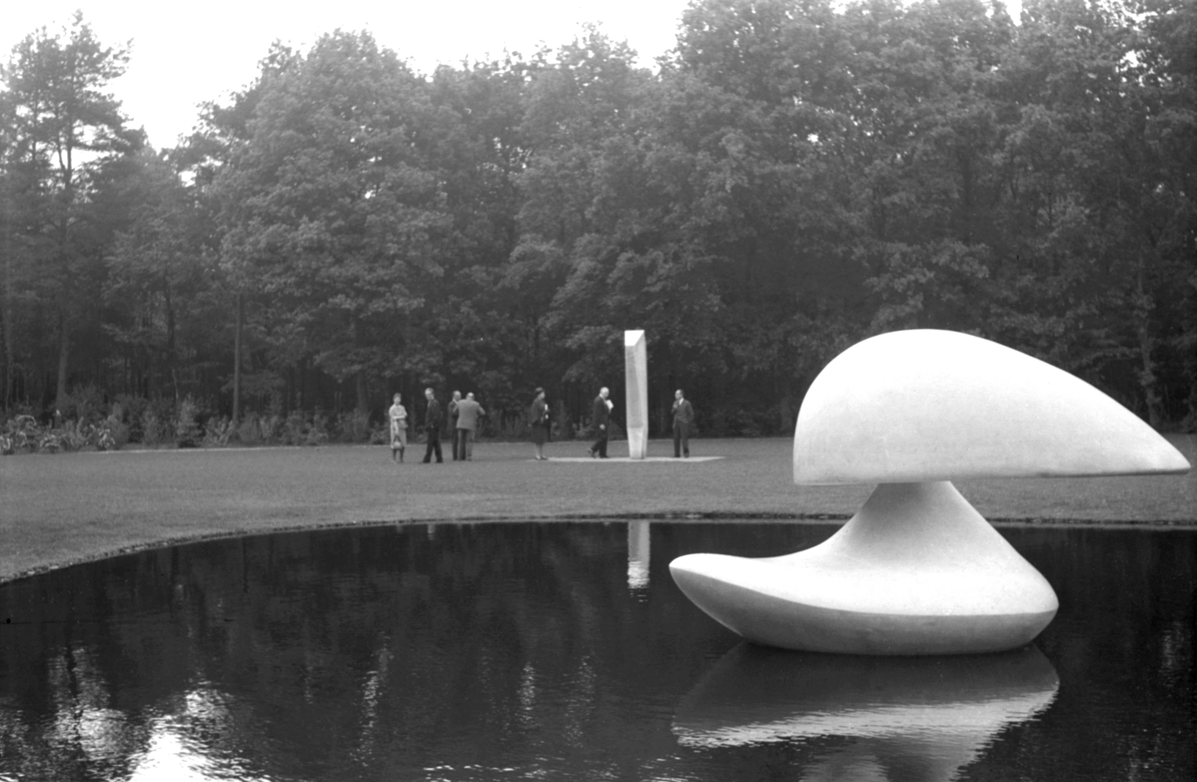 Opening beeldentuin, 3 juni 1961 Zwart-witfoto van de opening van de beeldentuin. Met een wit zwaanachtig beeld dat drijft in een vijver. En een groep mensen op een grasveld eromheen, bij een kunstwerk dat lijkt op een verticale zuil. Op de achtergrond staan bomen.