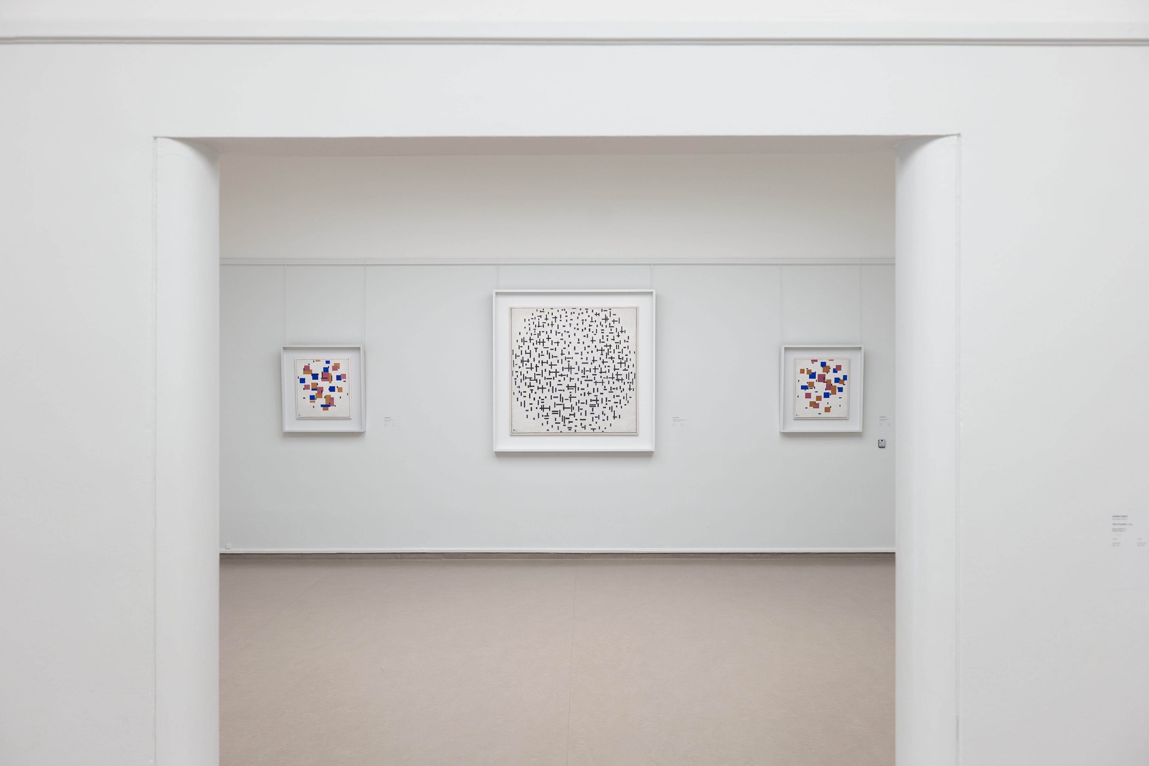 Foto eines Museumsraums mit drei abstrakten Gemälden an einer weißen Wand, wobei das größte Werk in der Mitte aus einem Muster aus schwarzen Punkten auf weißem Hintergrund besteht und die beiden kleineren Werke an den Seiten farbenfrohe geometrische Formen zeigen.