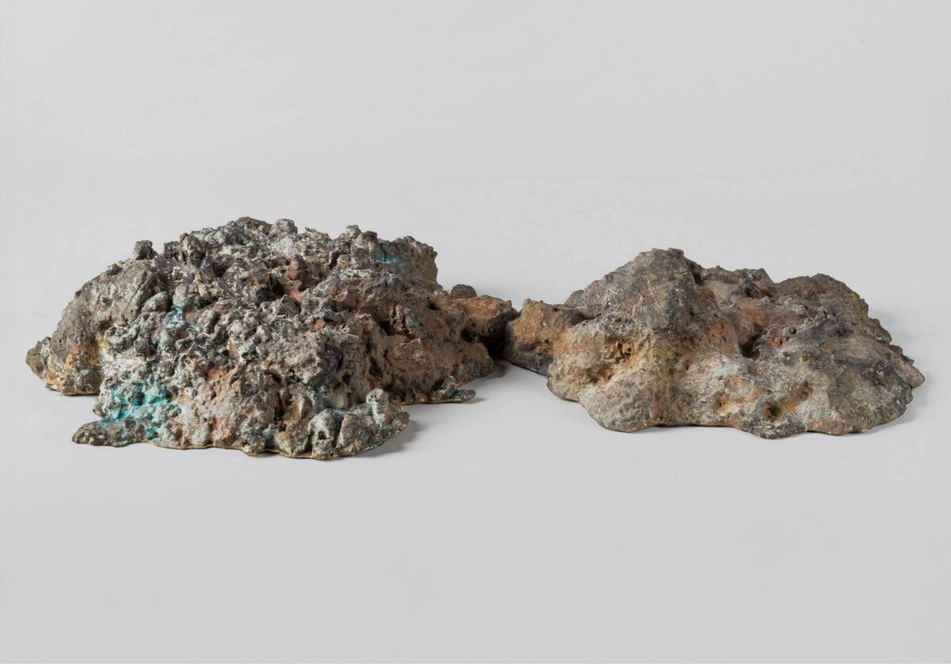 Sarah van Sonsbeeck, _Moles of Modernism - 00_003_, 2023 Twee ruwe, rotsachtige sculpturen in grijsbruine tinten, naast elkaar geplaatst op een lichte achtergrond, met een korrelige textuur en subtiele groene verkleuringen.