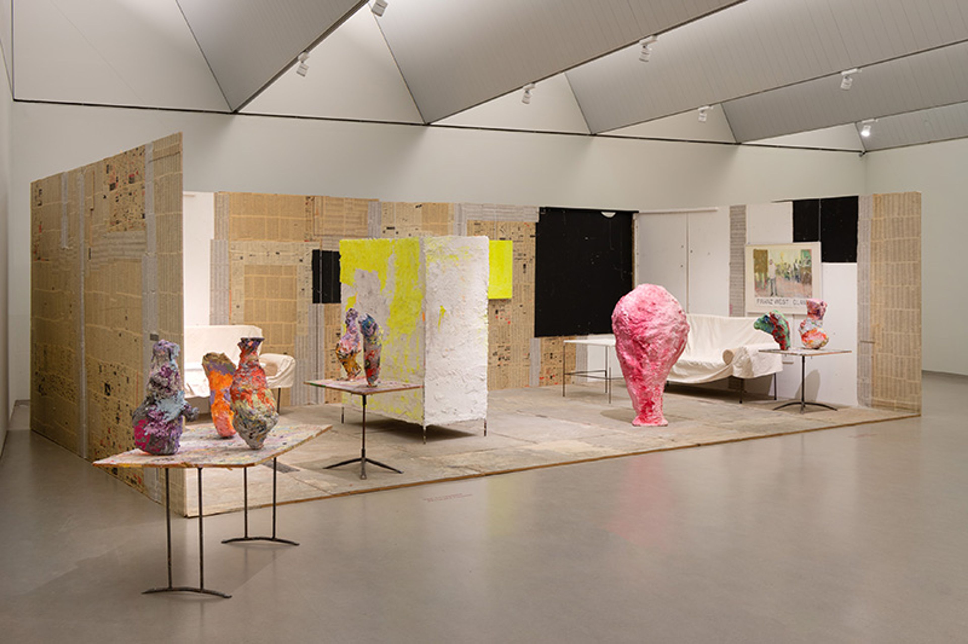 Franz West, Clamp, 1995 Foto van een kunstinstallatie met abstracte, kleurrijke sculpturen geplaatst op tafels en vloer. Ruimte bevat wanden met kartonachtige panelen en doeken met opvallende gele en zwarte vlakken