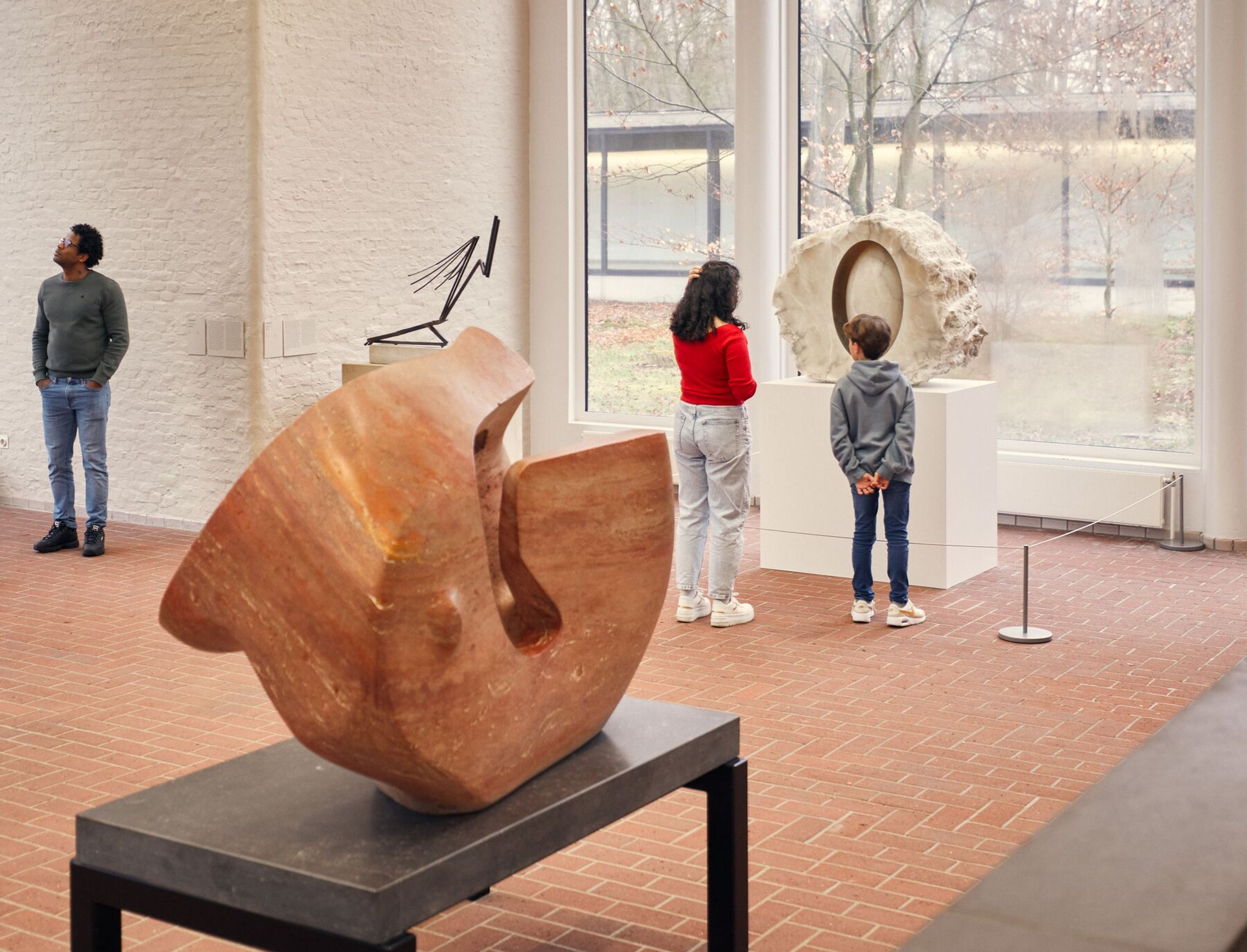 Foto van de beeldenzaal met drie bezoekers die verschillende beelden bekijken, waaronder een houten abstract beeld op een standaard en een klassieke naakte vrouwenfiguur op een sokkel. Grote ramen zorgen voor veel natuurlijk licht en laten de buitenomgeving met bomen zien. De vloer is van rode bakstenen gemaakt. 