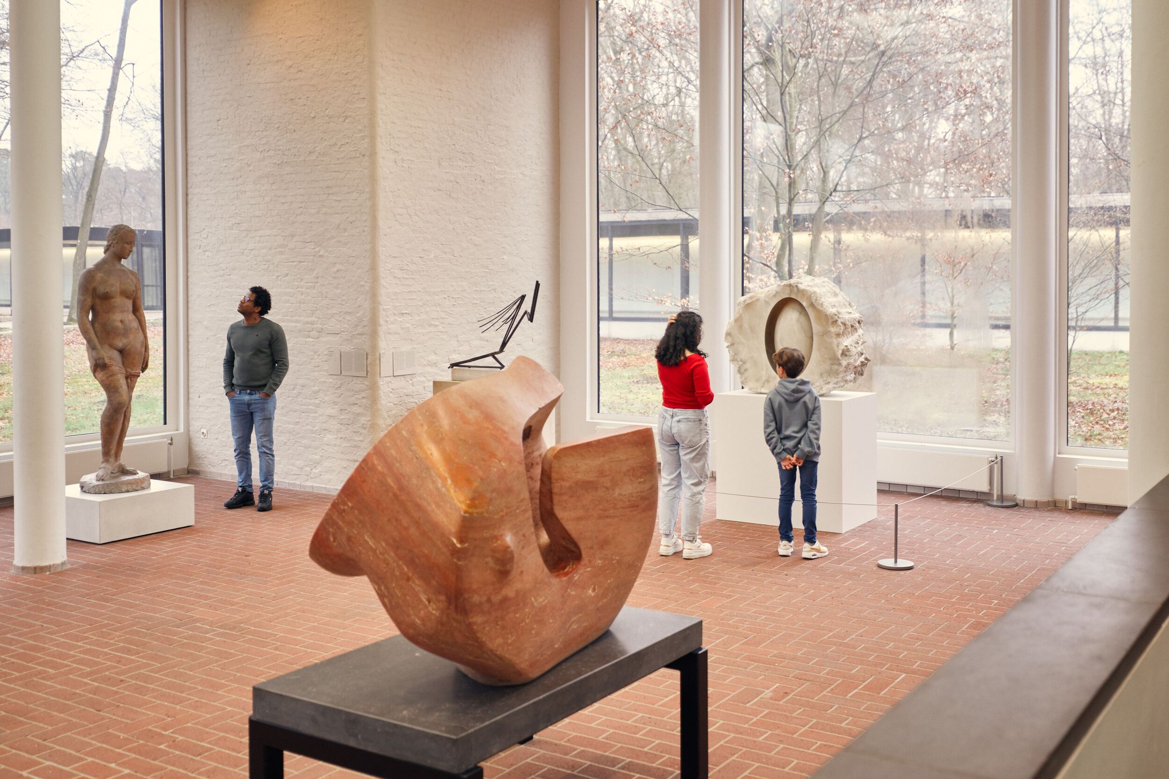 Foto des Skulpturensaals mit drei Besucherinnen und Besuchern, die verschiedene Skulpturen betrachten, darunter eine abstrakte Holzskulptur auf einem Sockel sowie eine klassische Frauenstatue in Aktdarstellung auf einem Podest. Große Fenster sorgen für viel Tageslicht und geben den Blick auf die Außenanlage mit Bäumen frei. Der Boden besteht aus roten Ziegelsteinen.