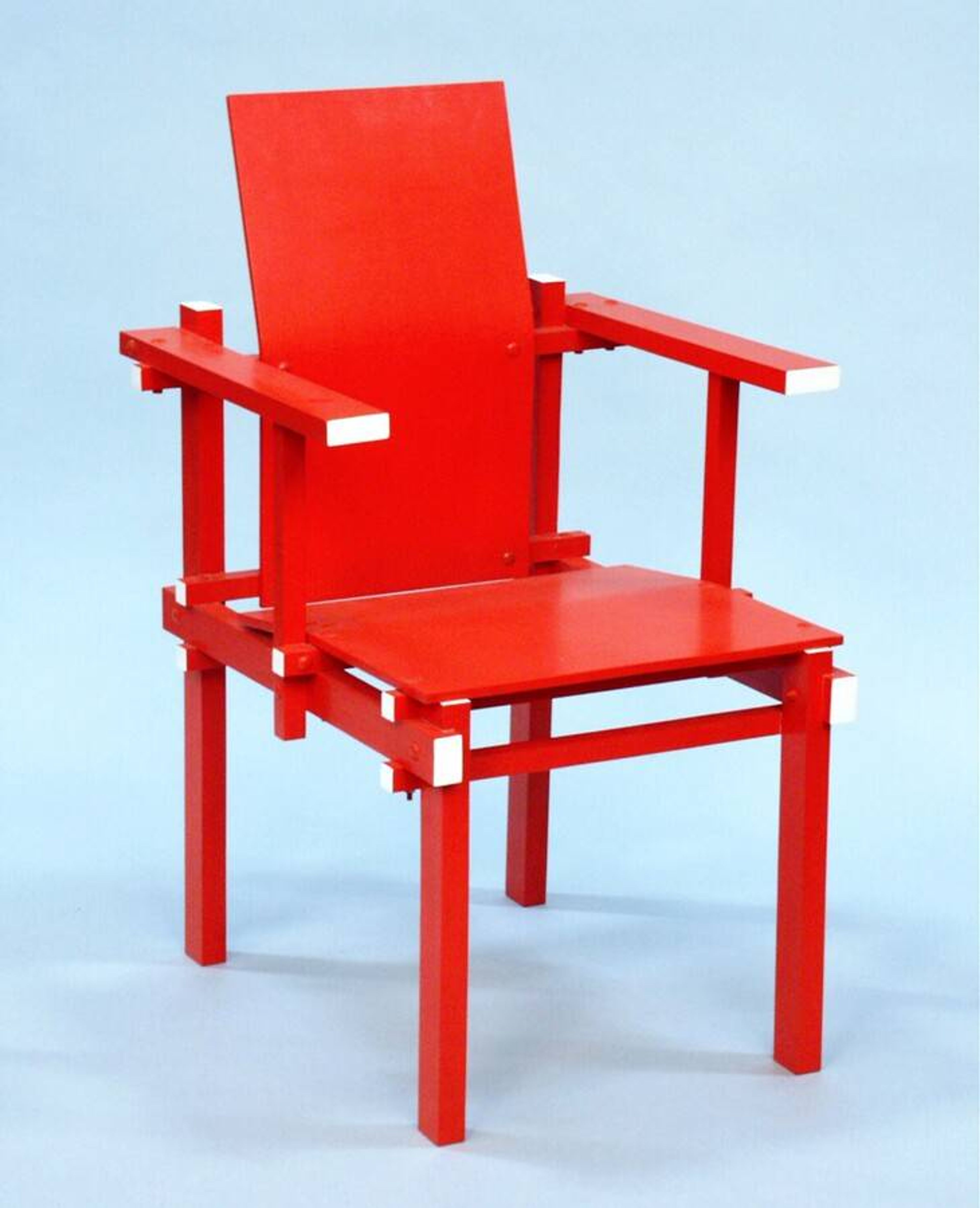 Gerrit Rietveld, stoel, ontwerp 1925, latere uitvoering Foto van een rode houten stoel met rechte lijnen en witte accenten op hoeken. Stoel heeft een hoge rugleuning, brede armleuningen en een minimalistische, geometrische stijl.