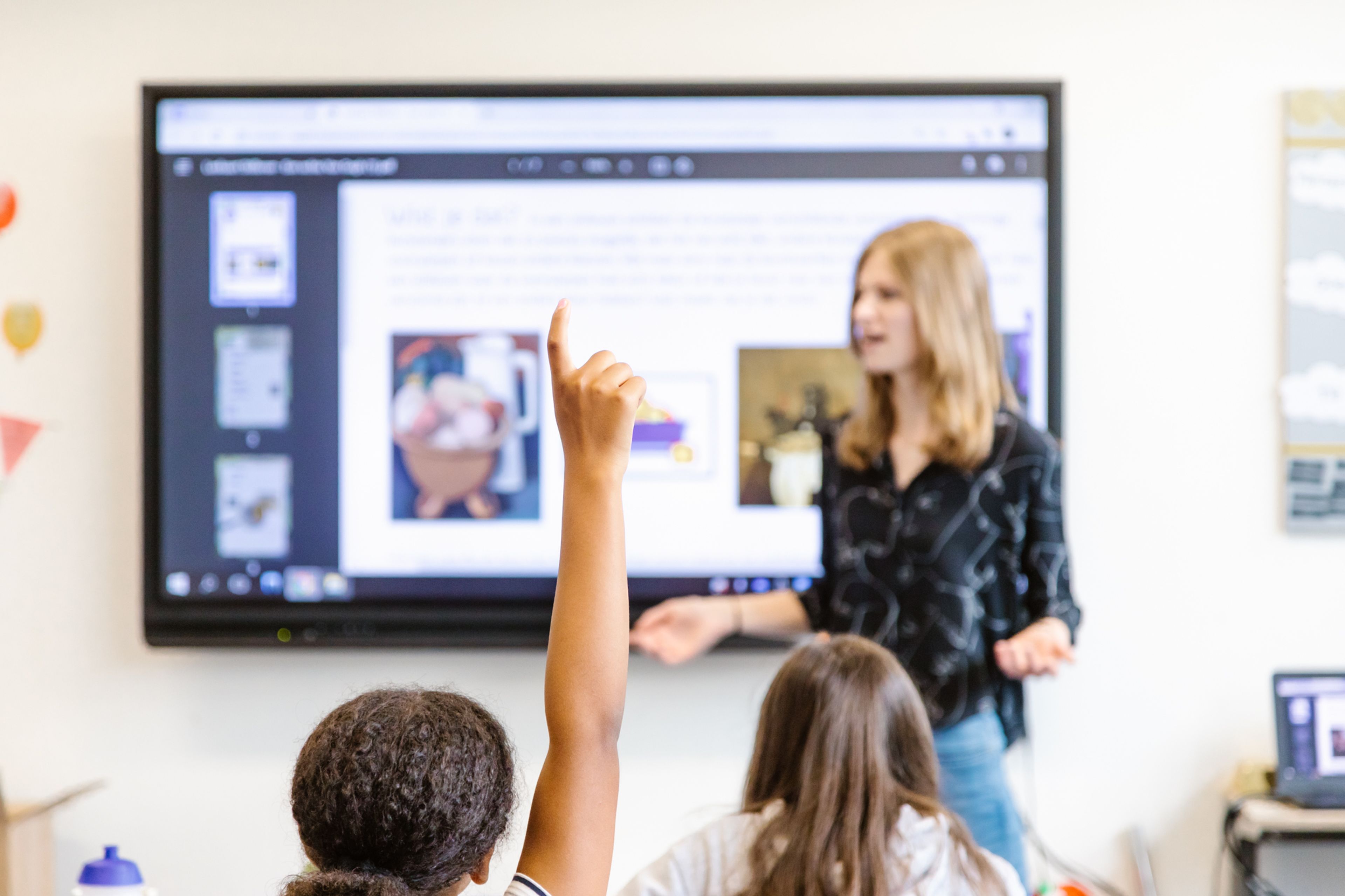 Foto van een klaslokaal. Op de voorgrond twee leerlingen, een leerling steekt hand op om een vraag te stellen. Op de achtergrond een docent voor een digibord